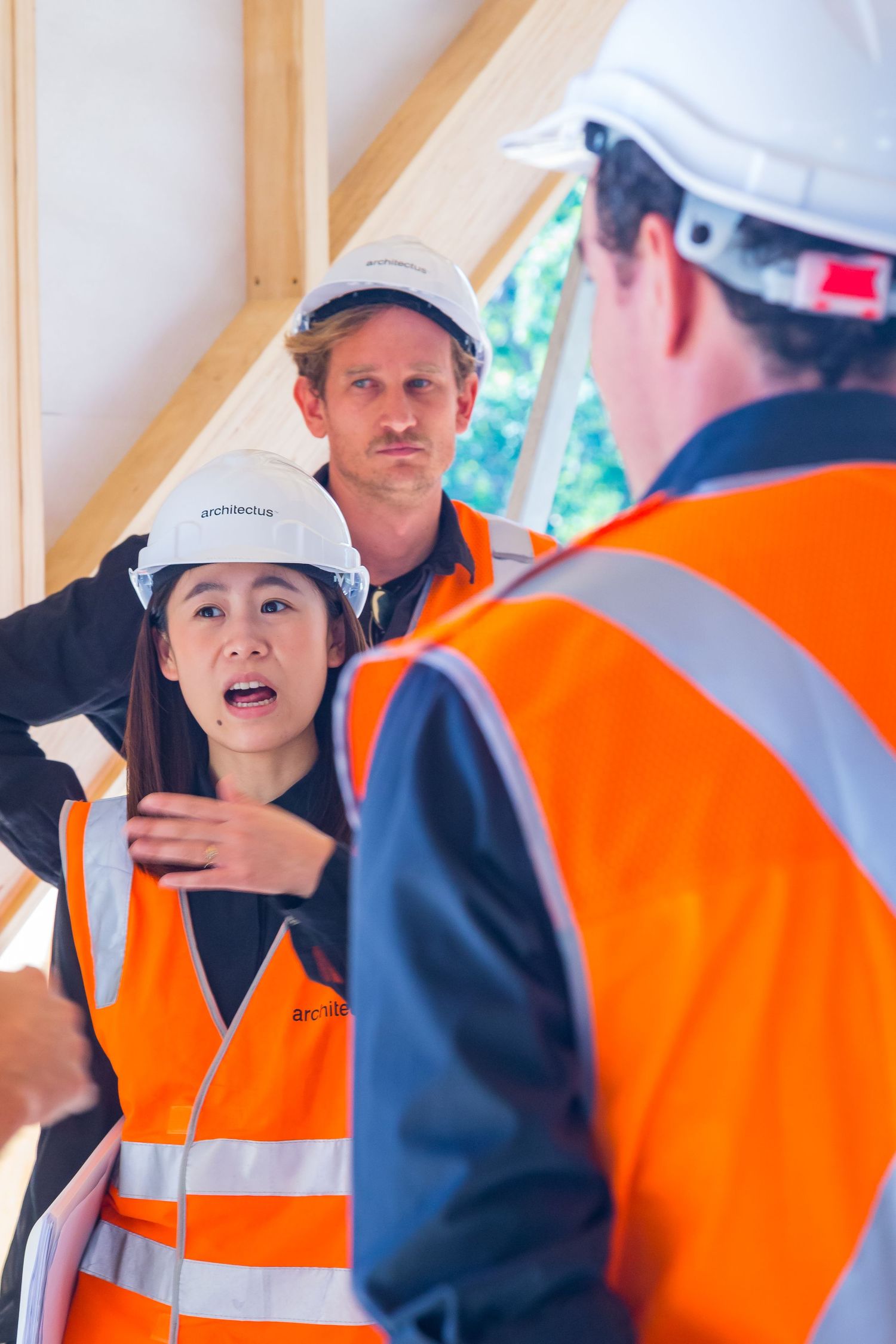 Construction workers in white hard hats and orange vests talk inside a timber-framed building while a woman gestures as she speaks.