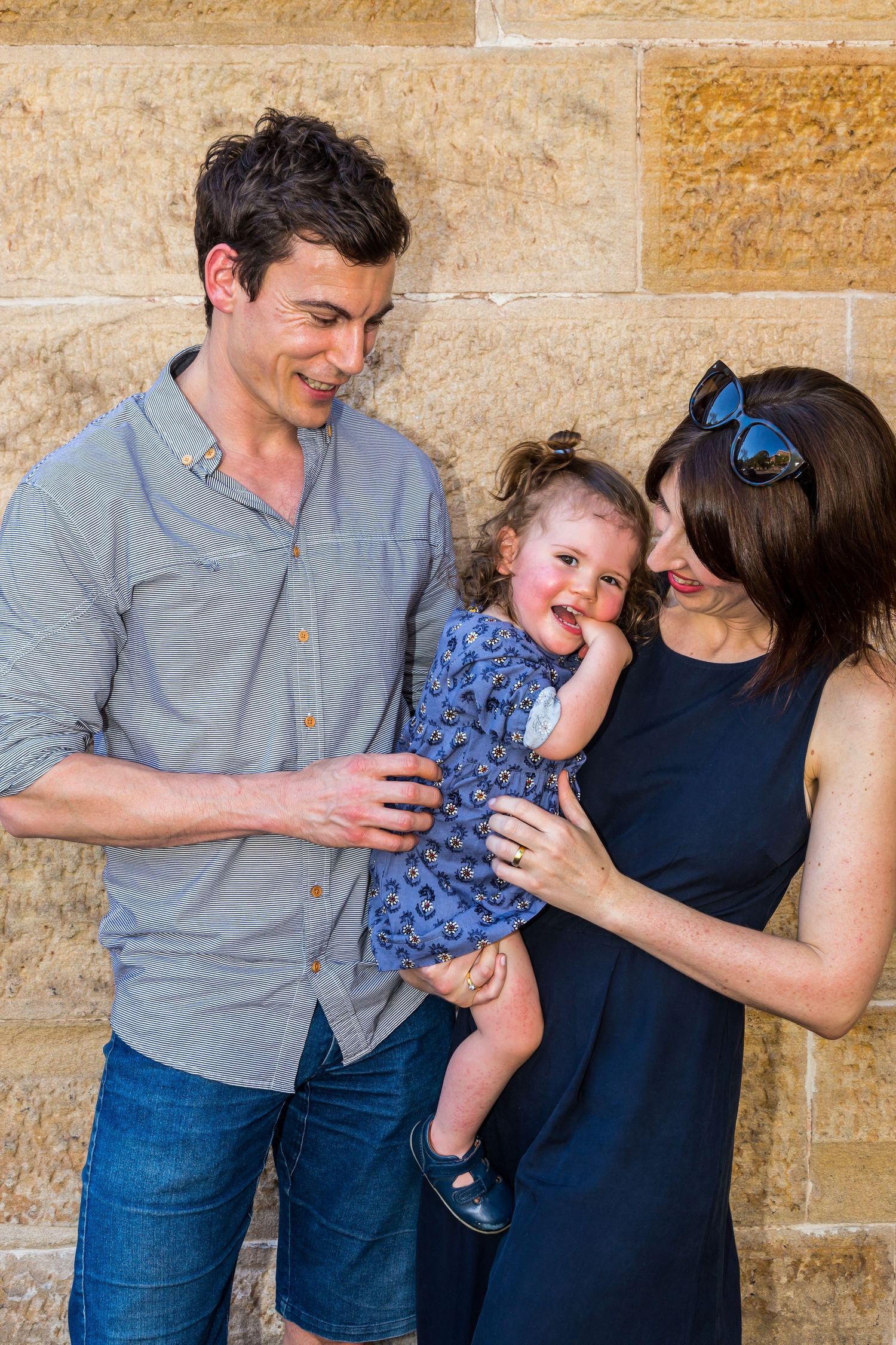 A smiling couple holds a laughing toddler in a blue patterned dress against a sandstone wall.