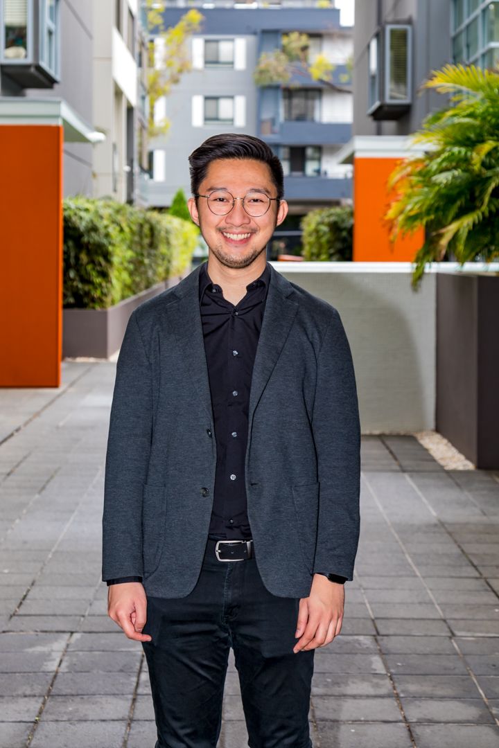Smiling man with glasses wearing a gray blazer and black shirt standing in a modern courtyard with orange accents and green plants.