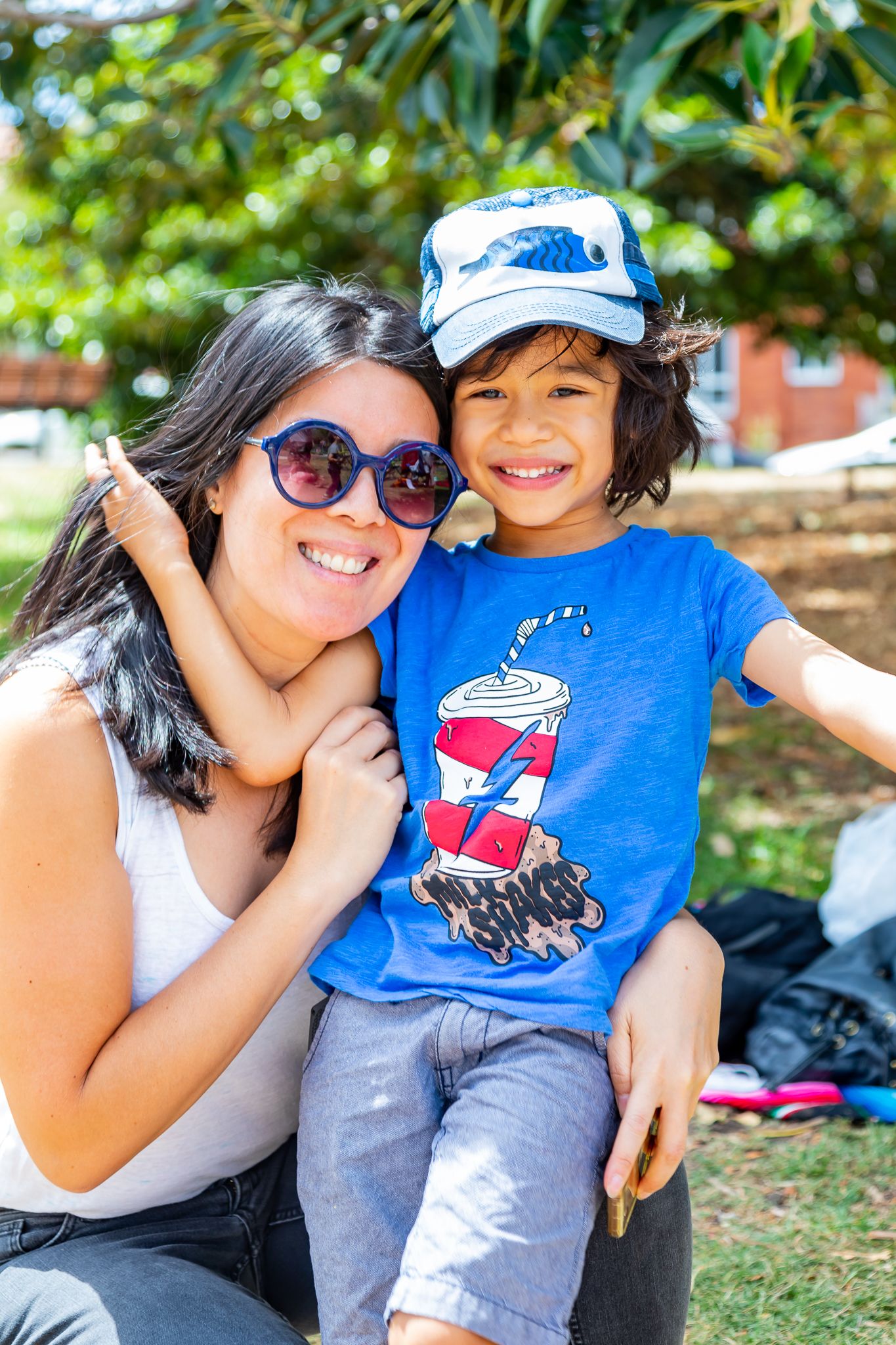 A smiling woman in round sunglasses hugs a young boy in a blue cap and graphic t-shirt in a sunny park.