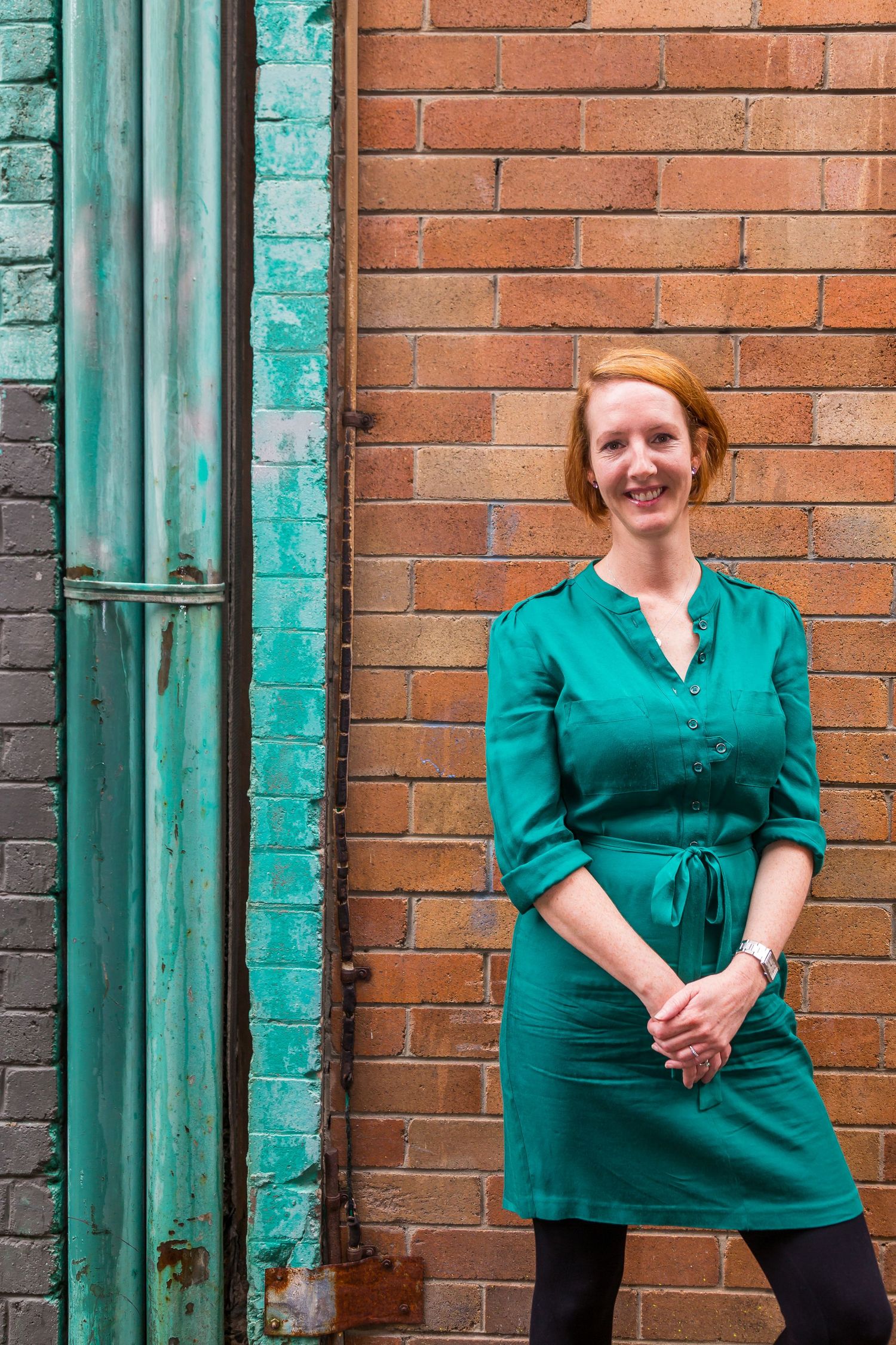 Red-haired woman in a green shirt dress smiles with clasped hands while standing against a rustic red brick wall with a green pipe.