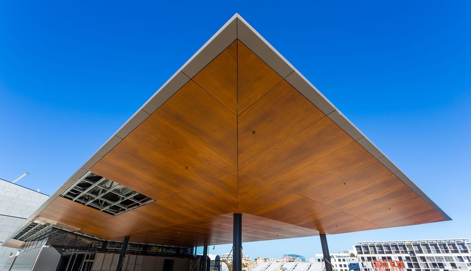 Wide upward view of a large timber-lined canopy roof with steel edges and a skylight panel against a deep blue sky.