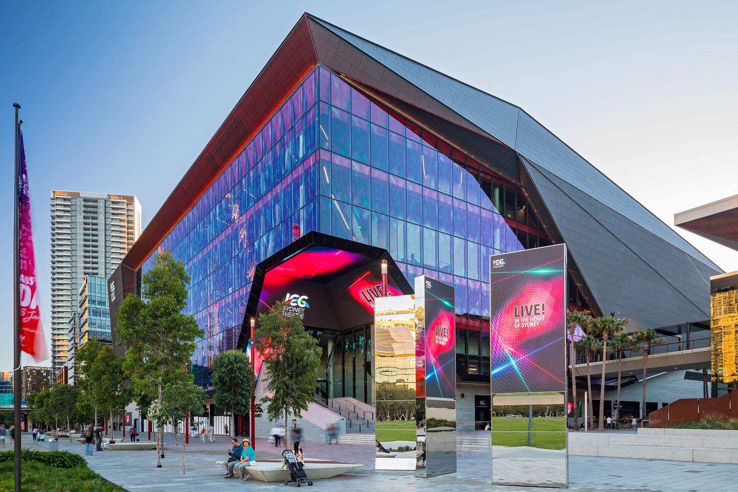 Angular glass-fronted entertainment venue with vibrant pink digital signage and tree-lined plaza under a clear blue sky.