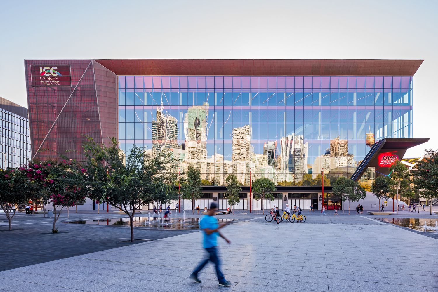 Glass-fronted convention centre reflecting city skyscrapers, with a red accent wall, trees and a blurred pedestrian crossing the plaza.
