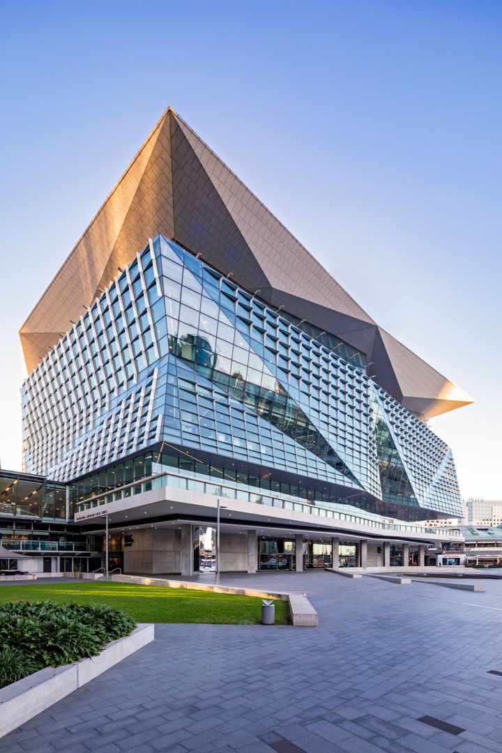 Striking angular convention centre with a pyramid-shaped bronze roof and faceted glass facade beside a green lawn.