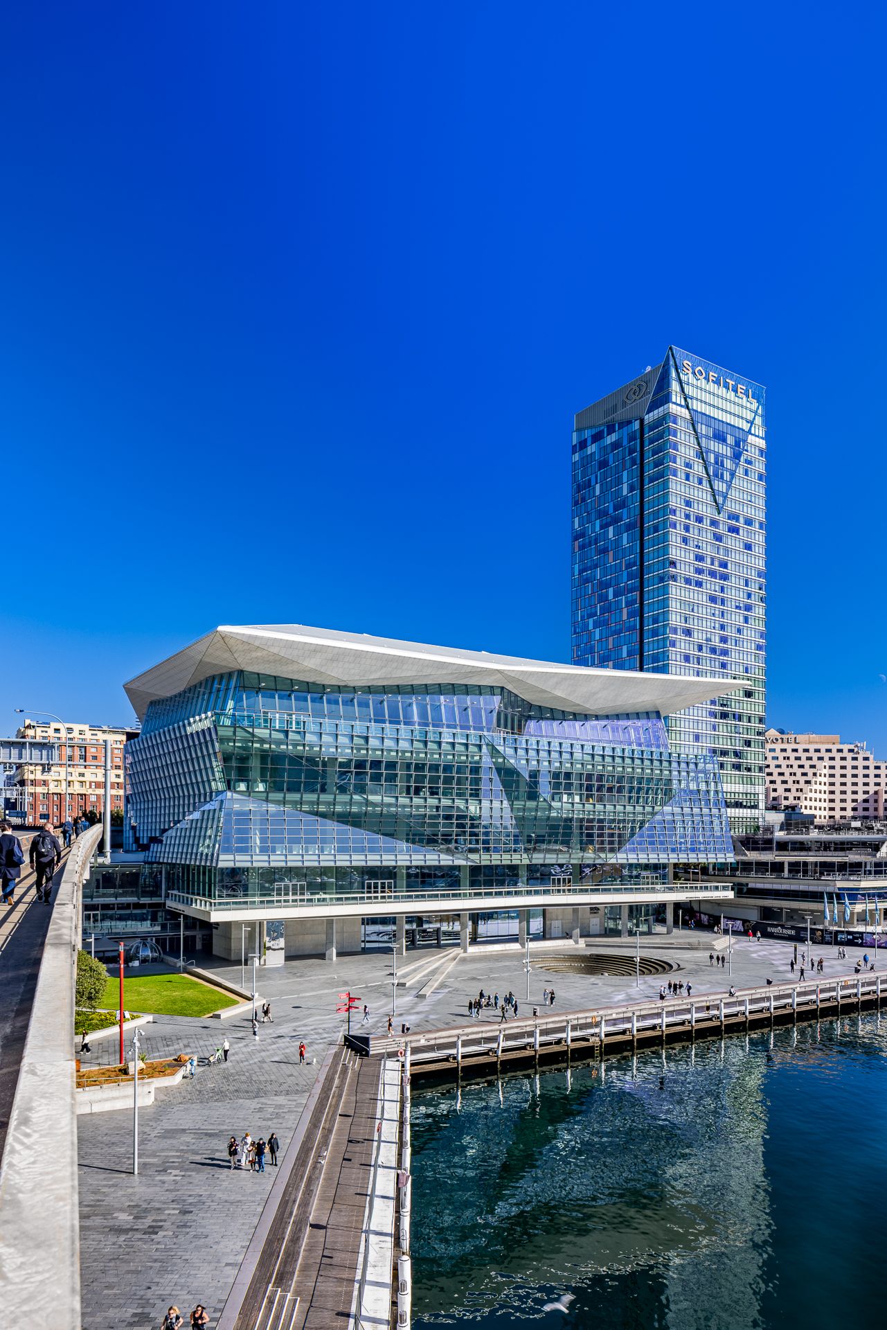 Angular glass convention centre with a sweeping white roof beside a tall blue skyscraper and a harbour waterfront walkway.
