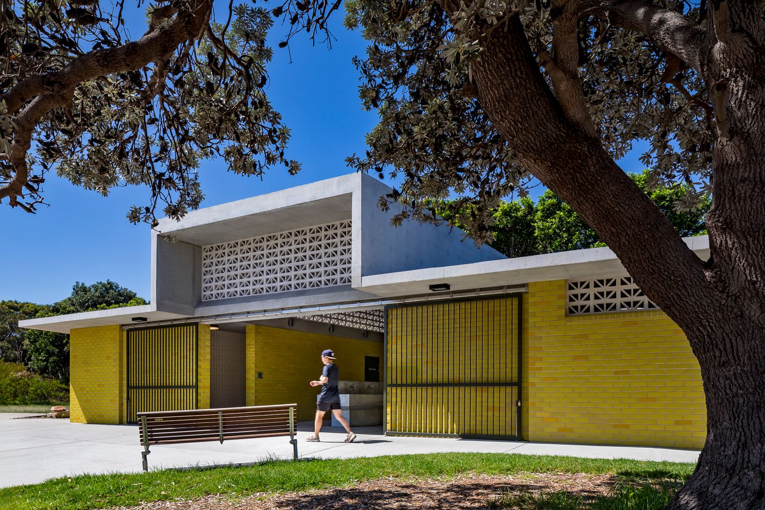 A pedestrian walks past a yellow brick amenities building with a decorative breeze block screen beneath a large shady tree.