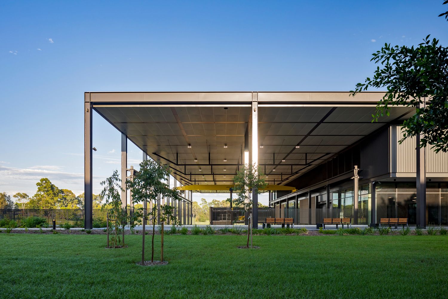 Wide steel canopy over an open covered area at a modern TAFE campus, with saplings and lawn under a deep blue evening sky.