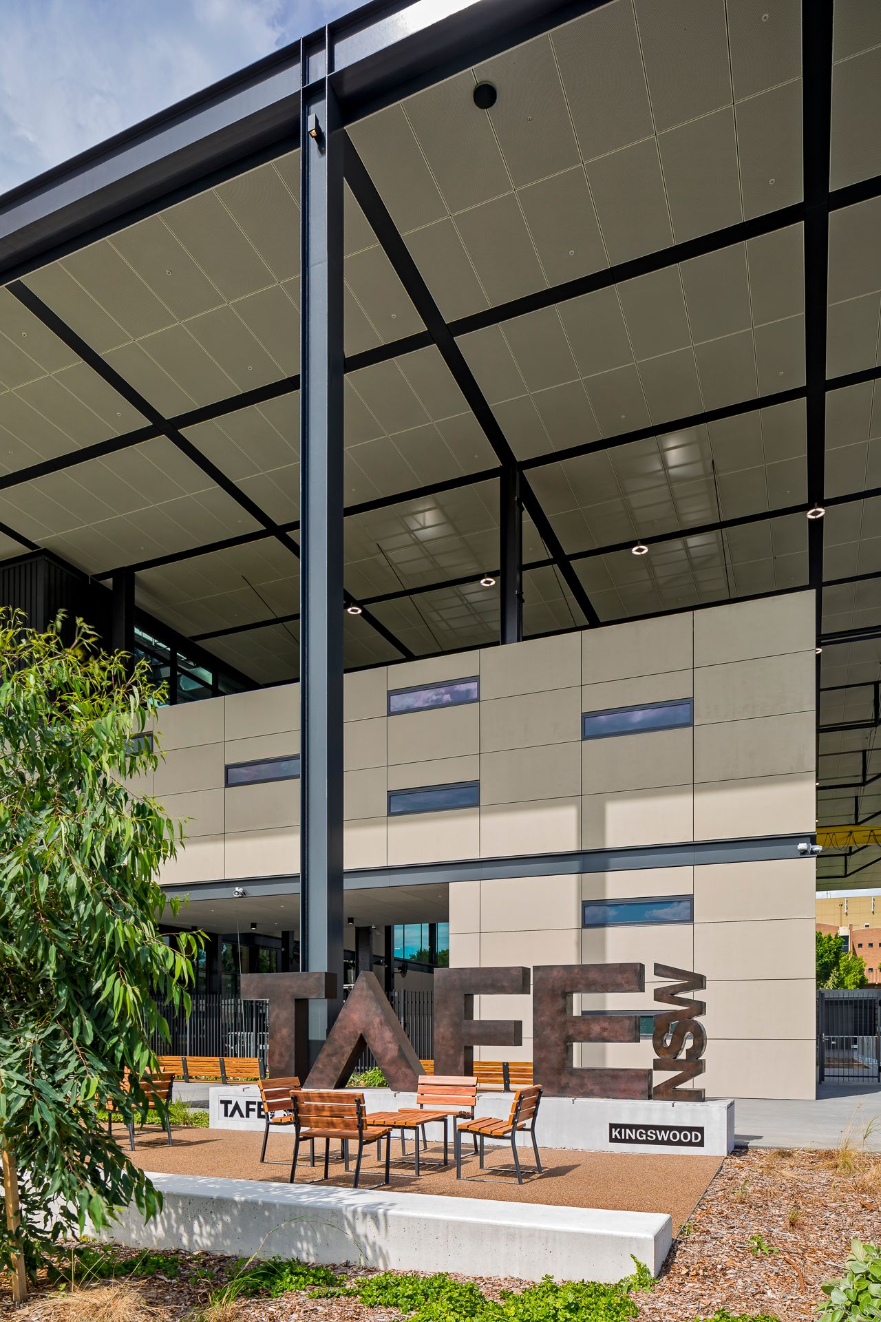 TAFE NSW Kingswood campus entrance with large steel lettering, outdoor timber seating and a soaring steel-framed canopy roof.