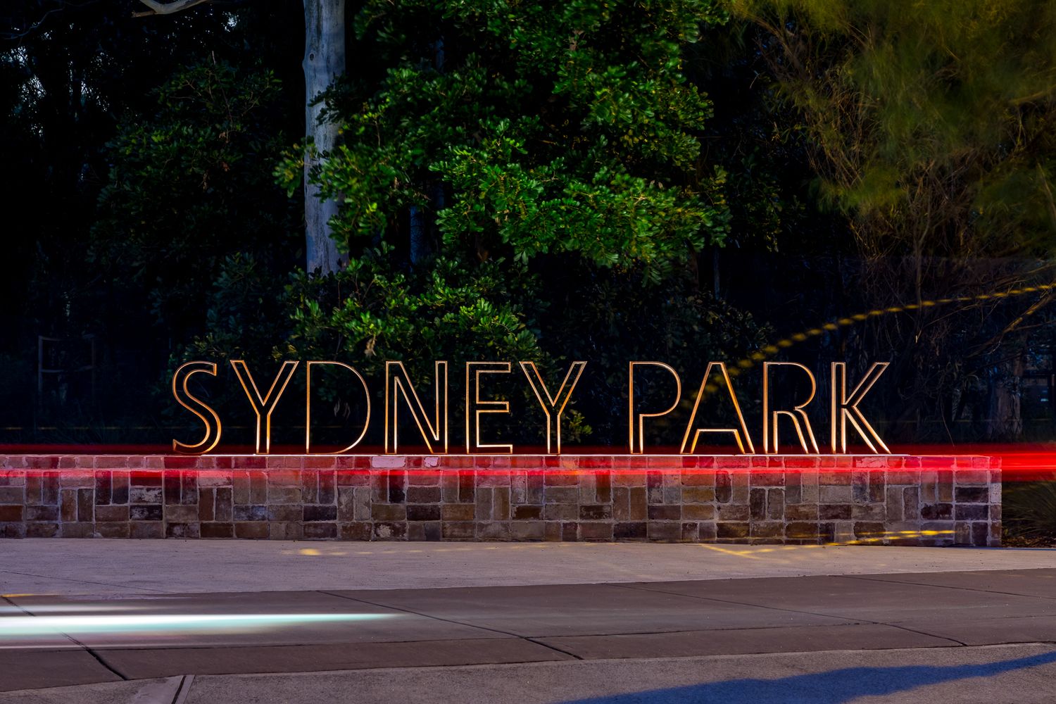 Illuminated Sydney Park sign glowing at night above a stone wall, with streaks of passing car lights in the foreground.