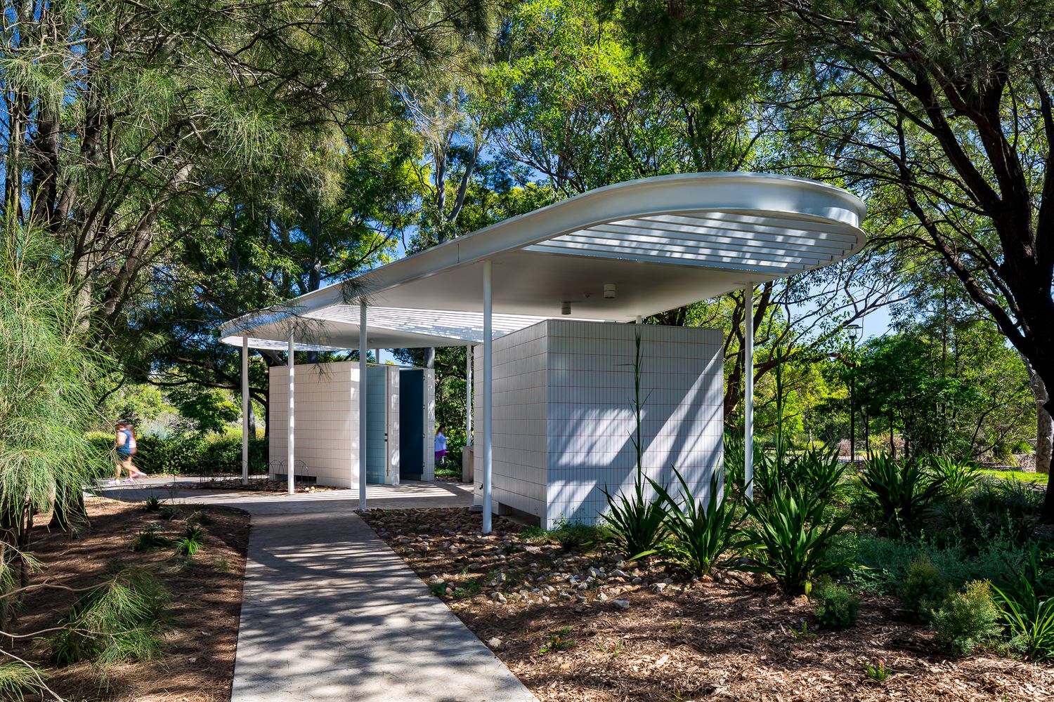 Curved white canopy roof shelters a tiled public amenities block with a stone path and spiky green plants in a leafy park.