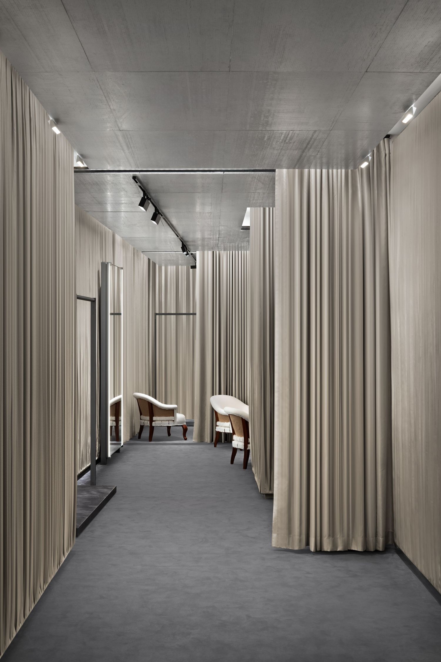 Fitting room corridor lined with floor-length taupe curtains, vintage armchairs, mirrors, and track lighting over gray carpet.