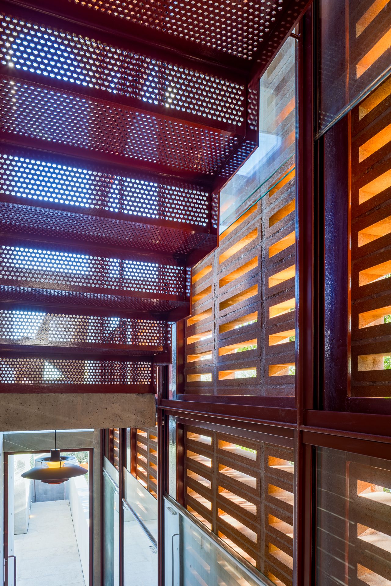 Perforated red metal stair and timber louvred screens casting warm light patterns, with a pendant lamp glowing below.