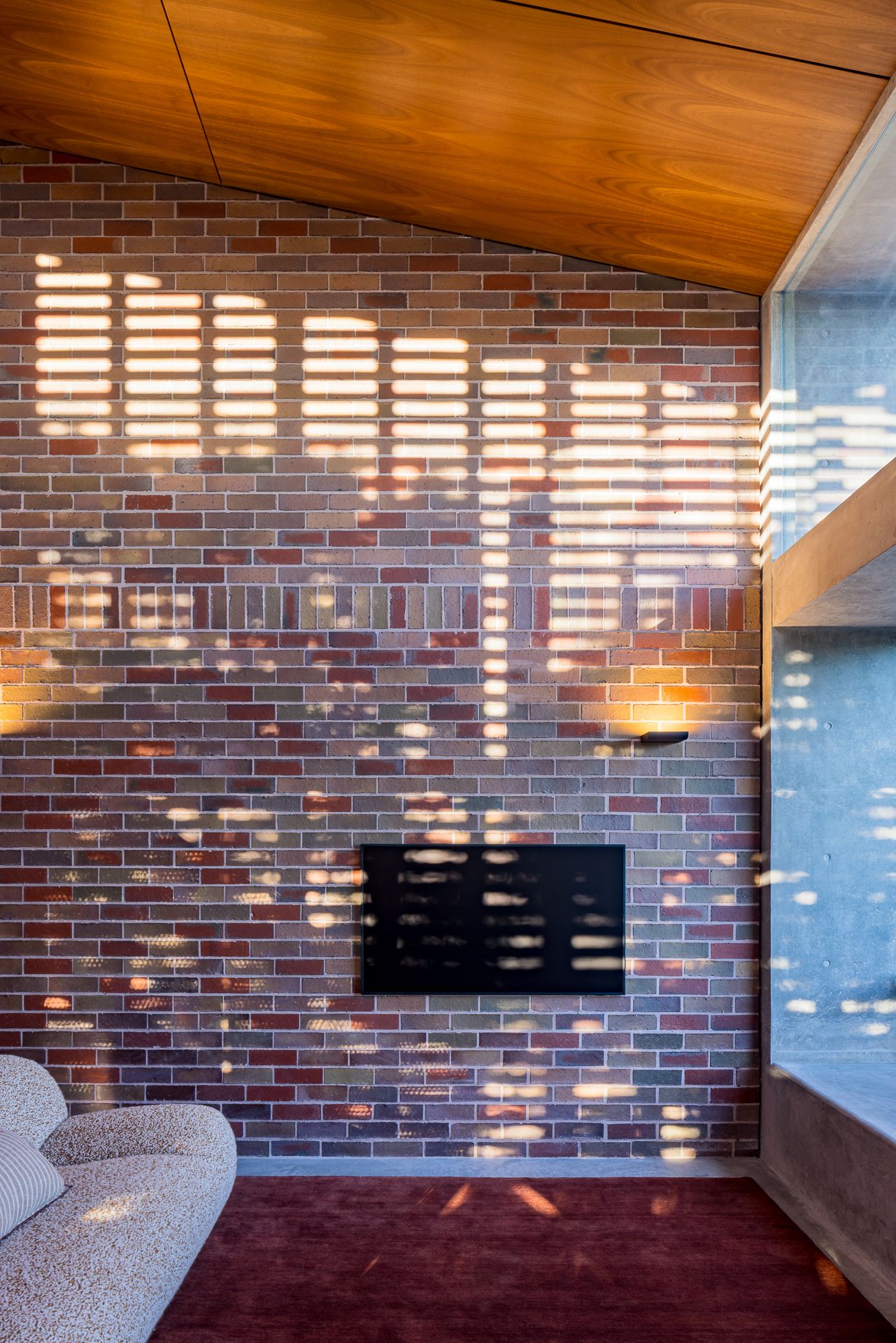 Living room brick feature wall with sunlight patterns, a timber ceiling, a wall-mounted TV and a textured cream sofa.