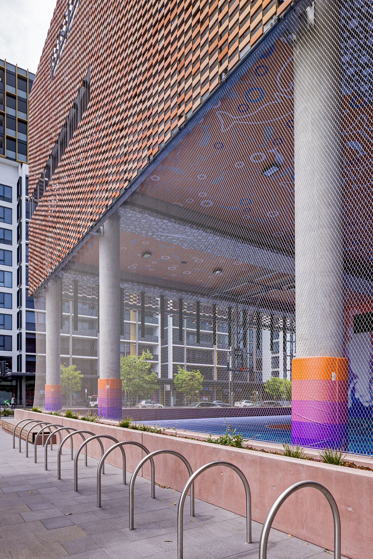 Modern building exterior with a perforated copper facade, round columns with colorful painted bases, bike racks and a reflective pool.