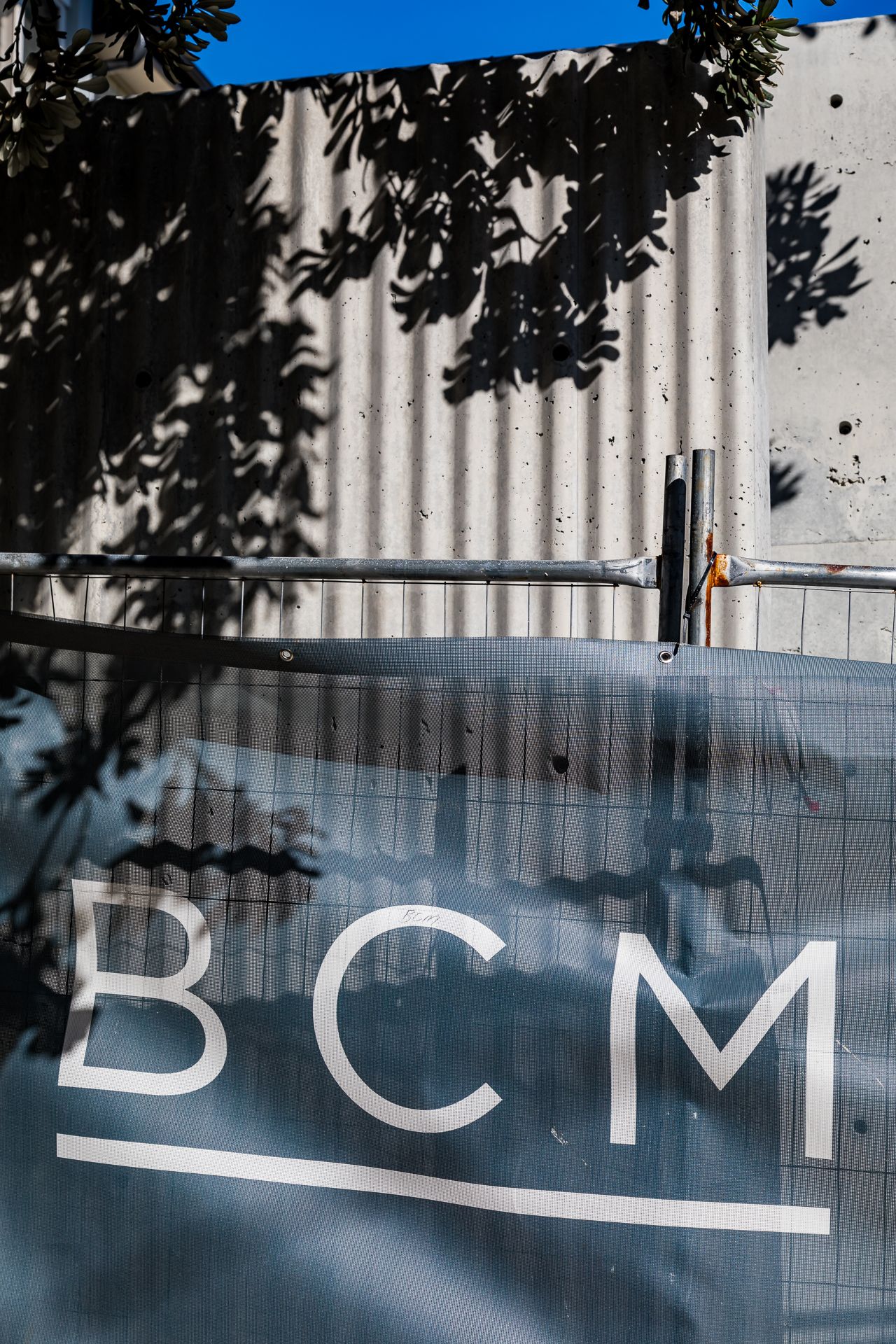 Corrugated concrete wall with tree shadows above a construction hoarding displaying the white BCM logo.