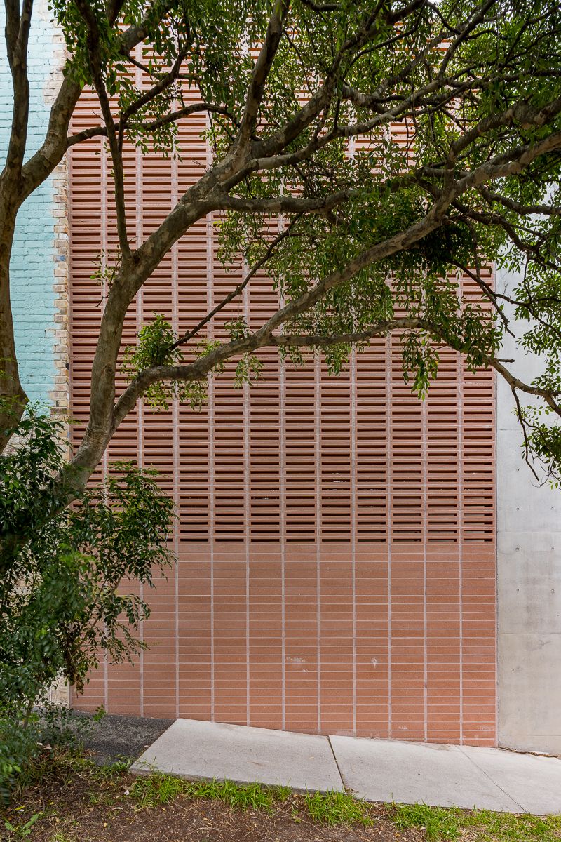 Terracotta louvered brick screen wall partly shaded by tree branches, set beside a concrete panel and a paved footpath.
