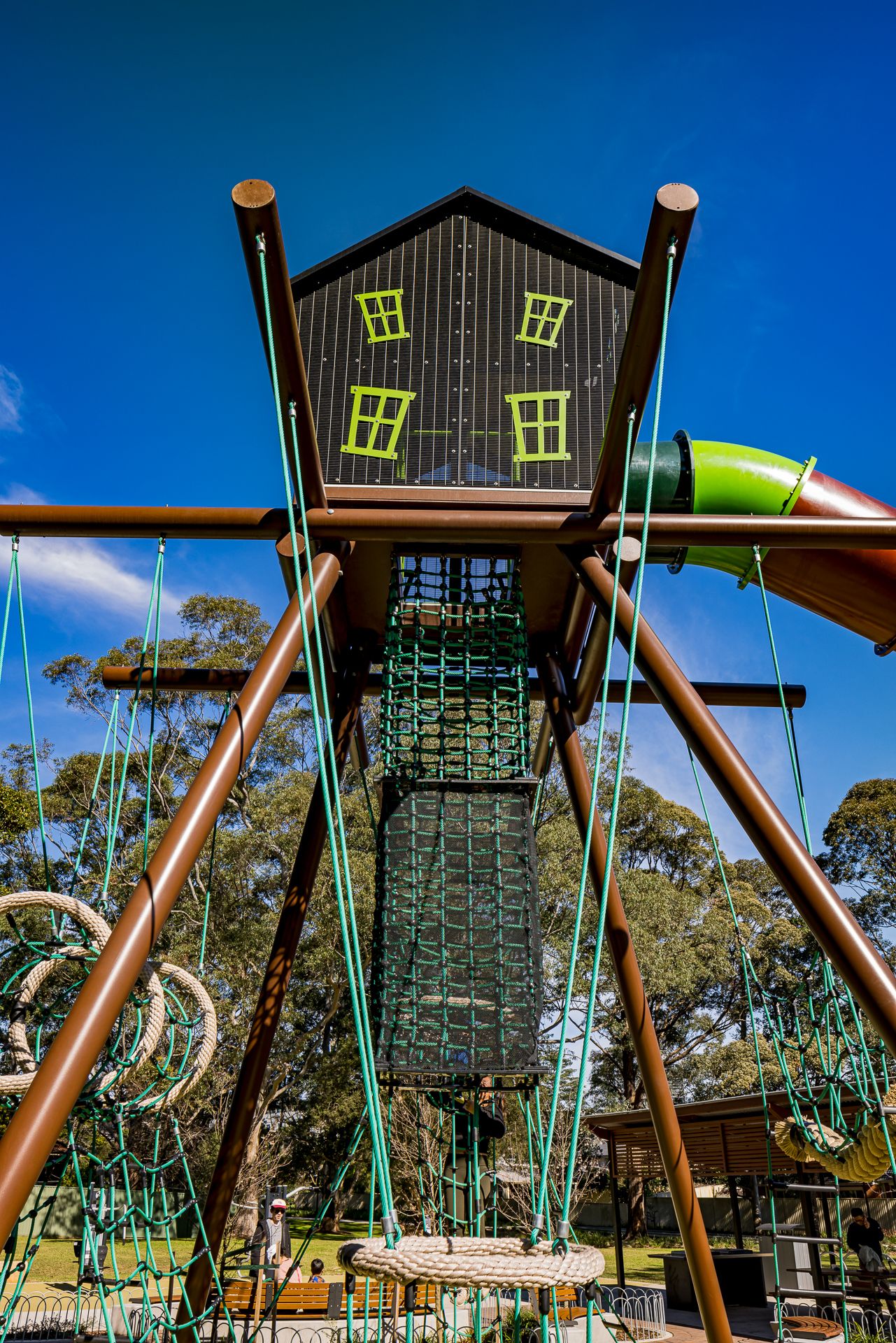 Close-up of a dark treehouse playground tower with green windows, climbing net, rope structures and slide against a deep blue sky.