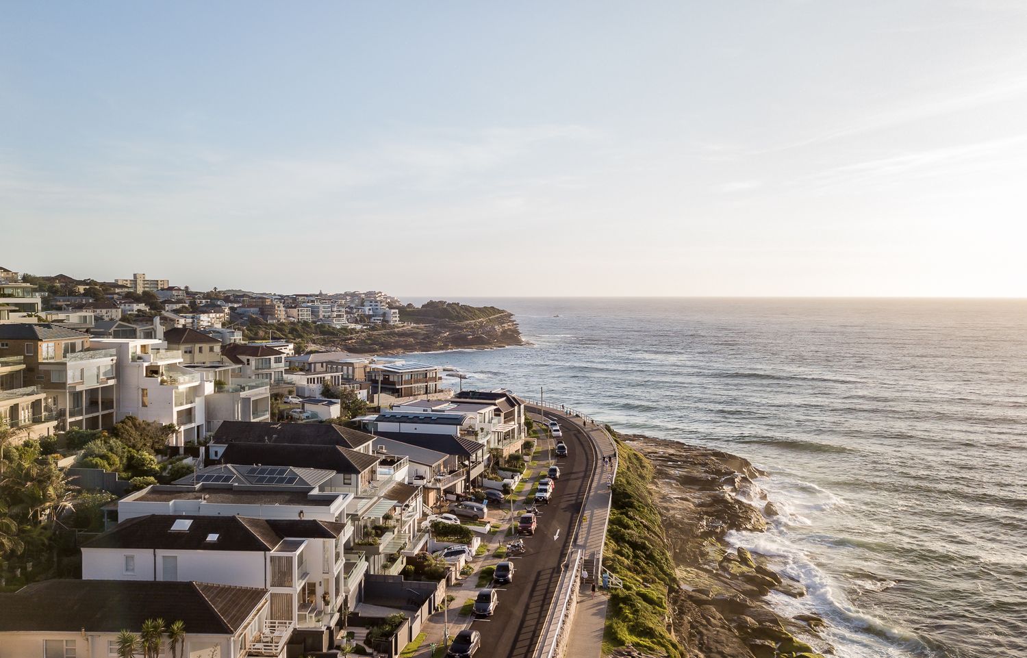 Aerial view of a coastal road lined with modern homes overlooking the ocean at sunrise.