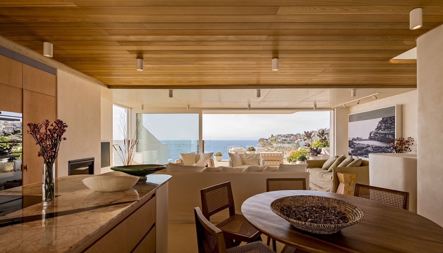 Open-plan kitchen and dining area with timber ceiling, stone benchtop and a living space opening to sweeping ocean views.