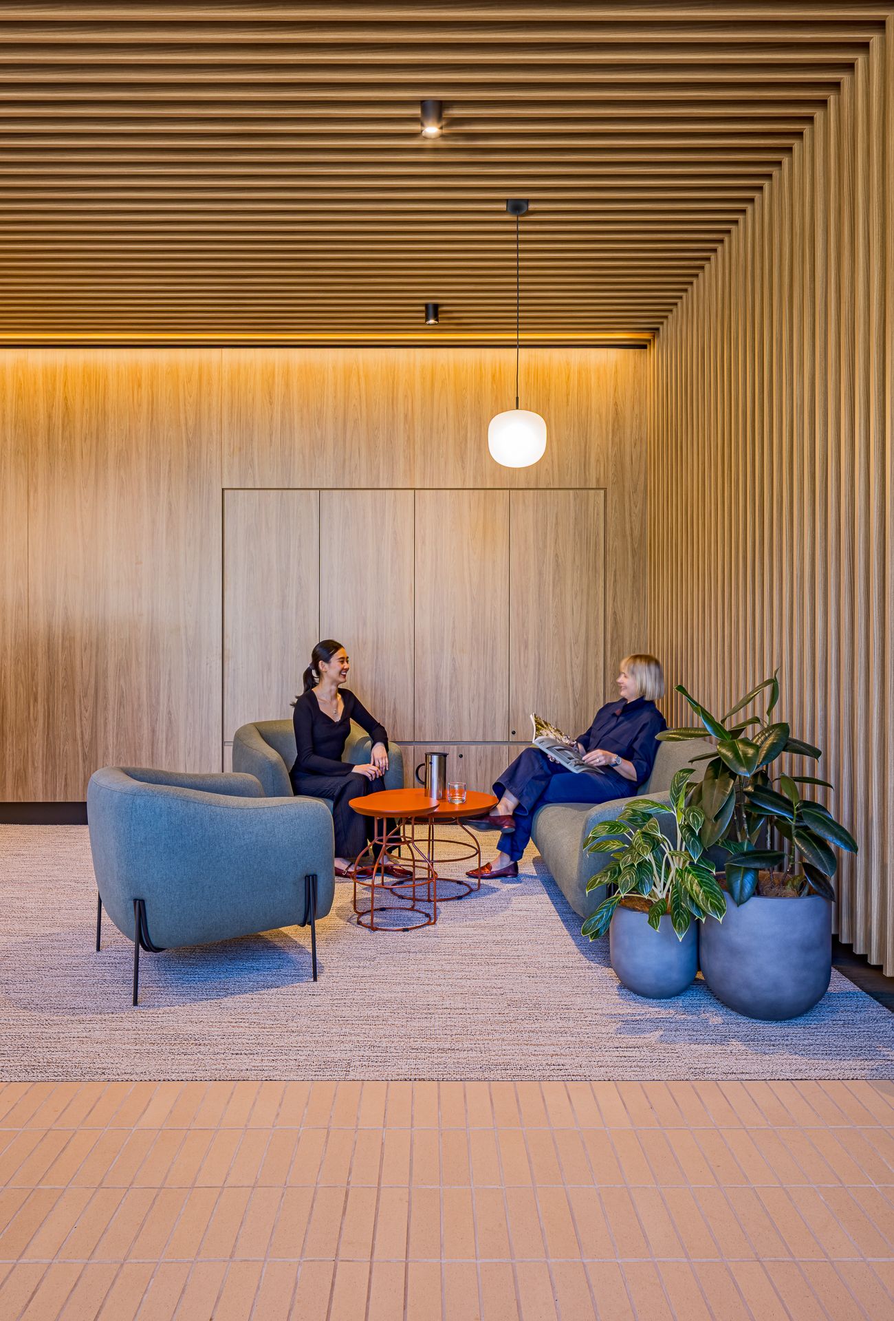 Two women chatting in a warm timber-lined lounge with blue-green armchairs, an orange coffee table and large potted plants.