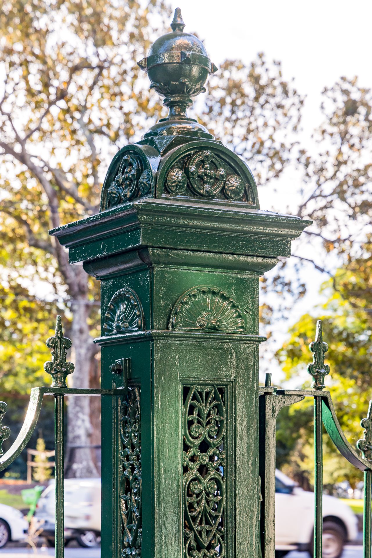 Detail of a green cast-iron gate post with decorative filigree panels and a domed finial against leafy trees.