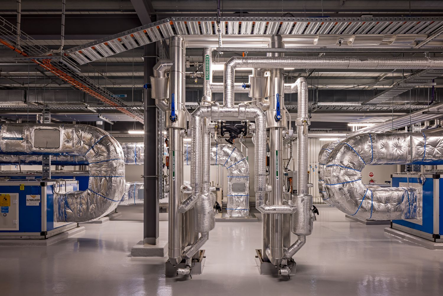 Industrial plant room with insulated stainless steel ductwork, large pipes and blue mechanical equipment on a polished concrete floor.