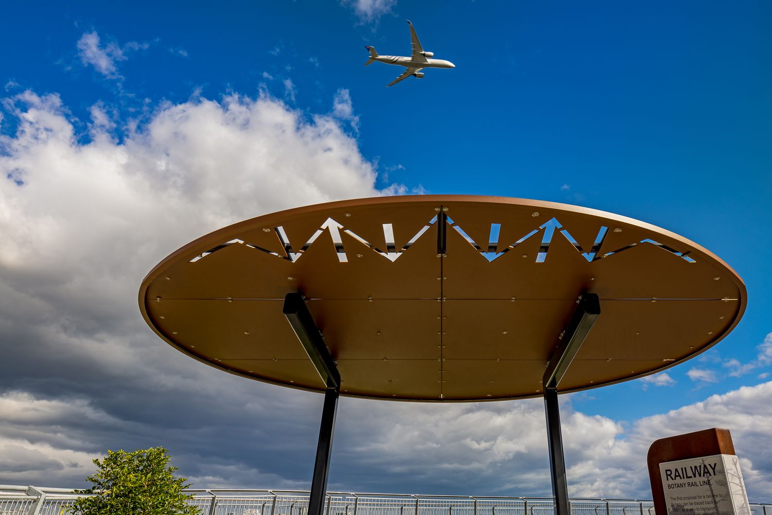 Round copper-toned shelter canopy with cut-out patterns on steel supports as an airplane flies through a cloudy blue sky above.