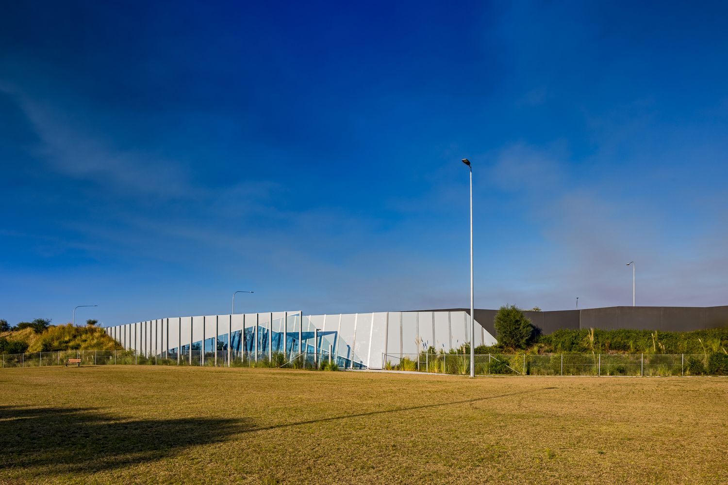 Modern glass-walled building with a long white facade beside a dry grassy field under a deep blue sky.