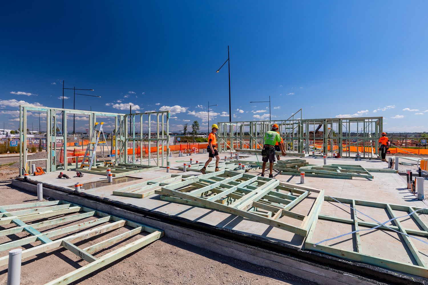 Construction workers in hard hats assemble green steel wall frames on a concrete slab at a residential building site.