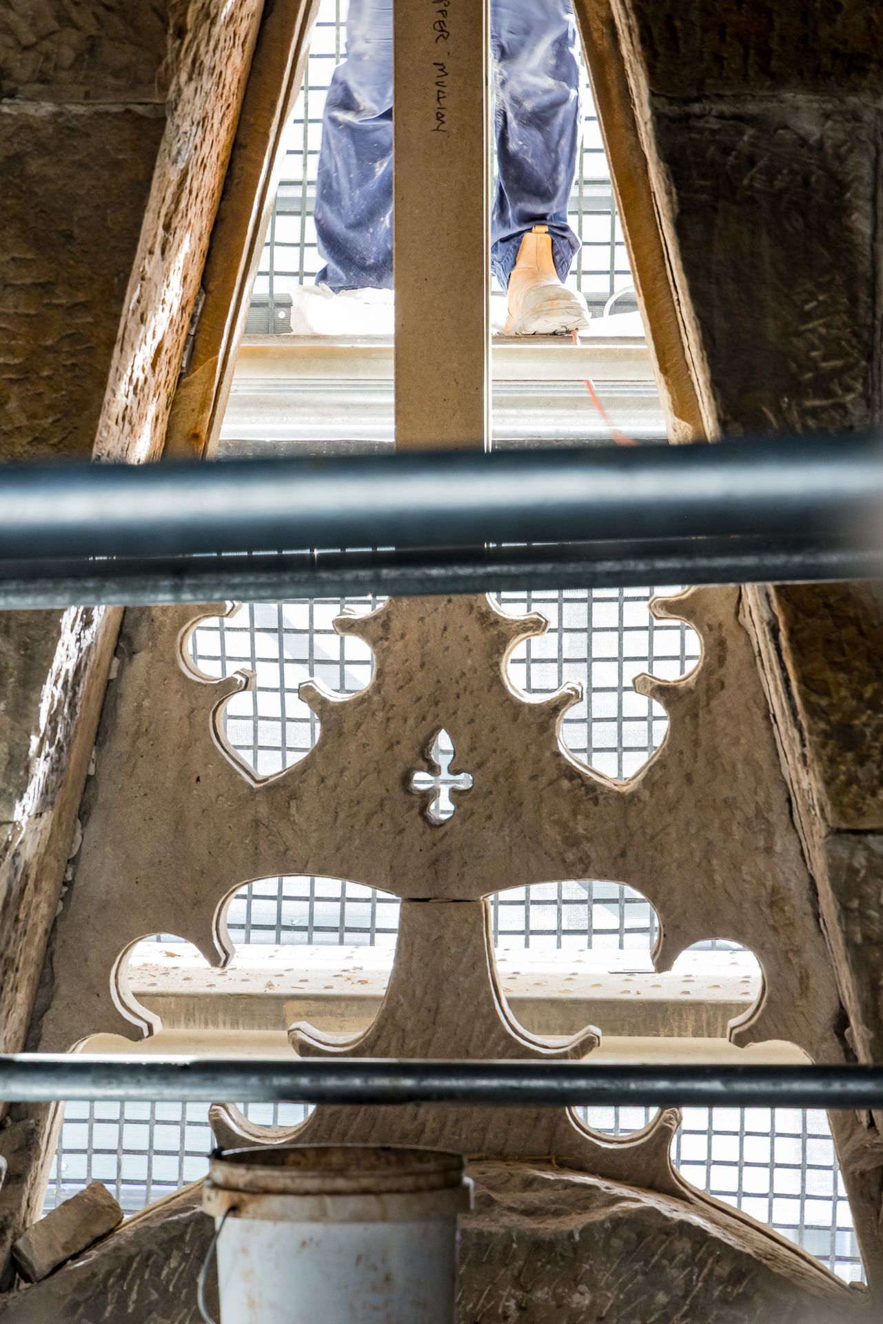 Gothic stone tracery with quatrefoil cutouts and a small cross, backed by scaffolding and a person standing above.