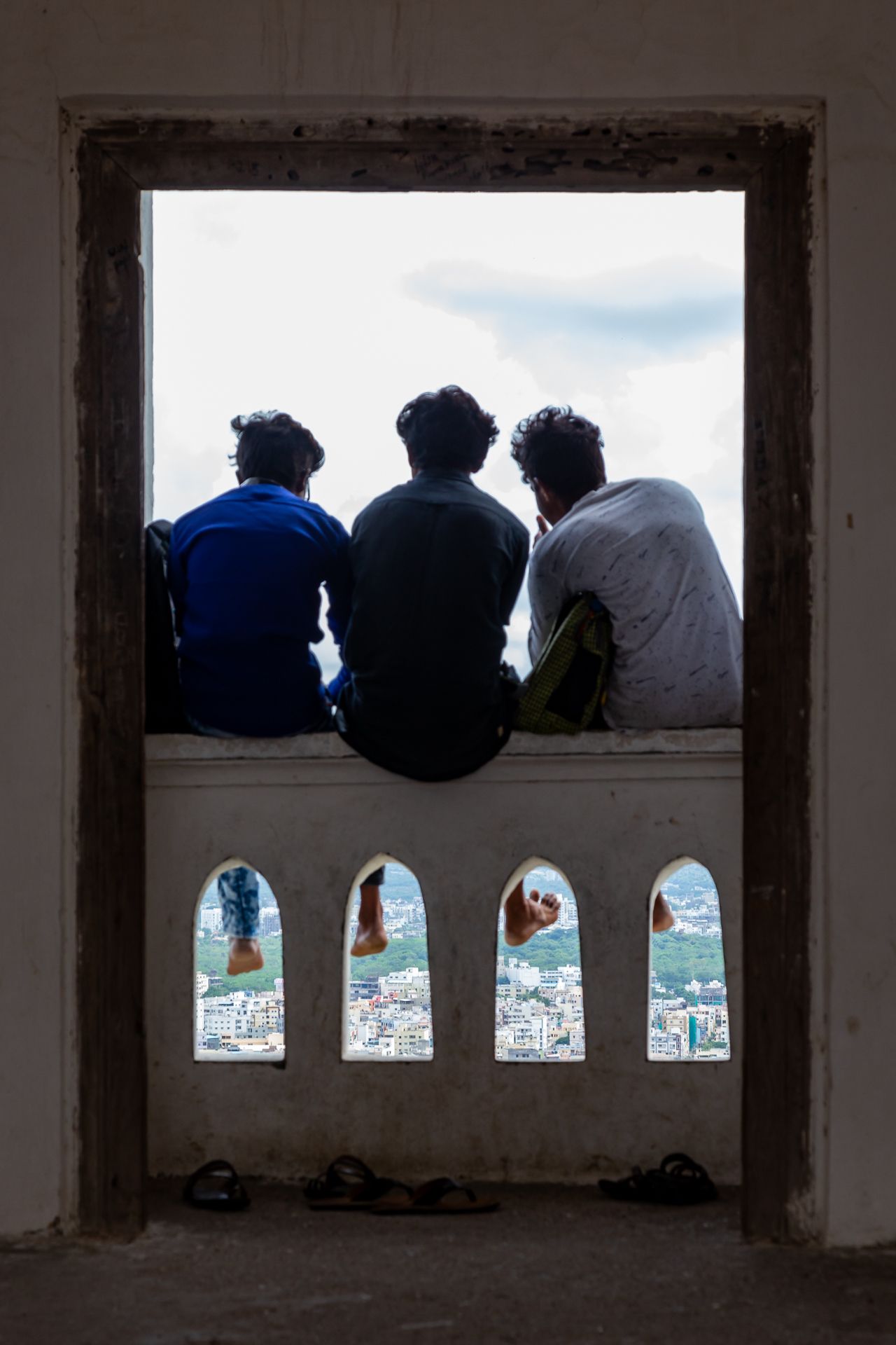 Three people sit on a ledge in a large stone window opening, looking out over a city skyline, with sandals on the floor below.