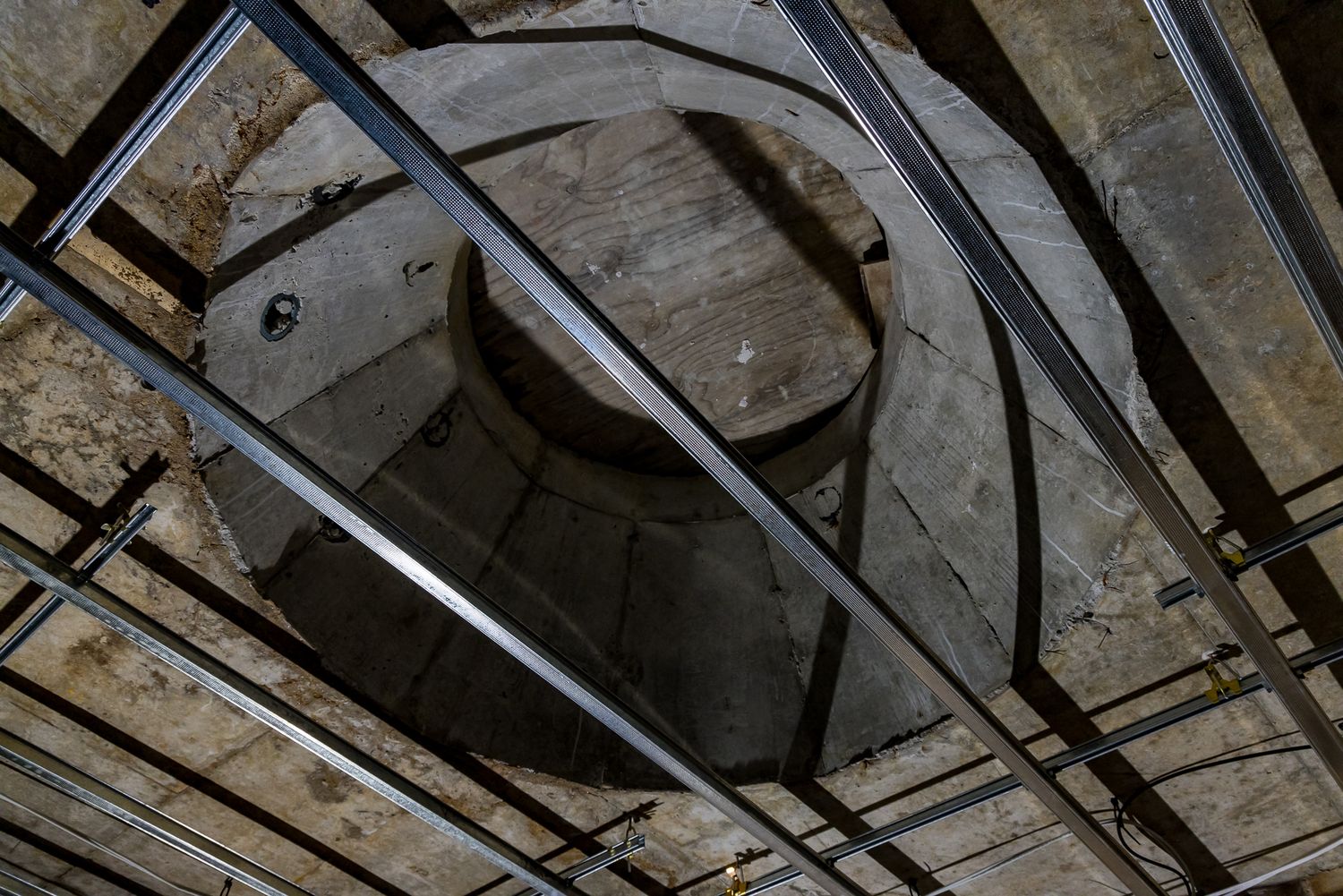 Upward view of a circular concrete opening in a ceiling crossed by metal framing and steel battens during construction.