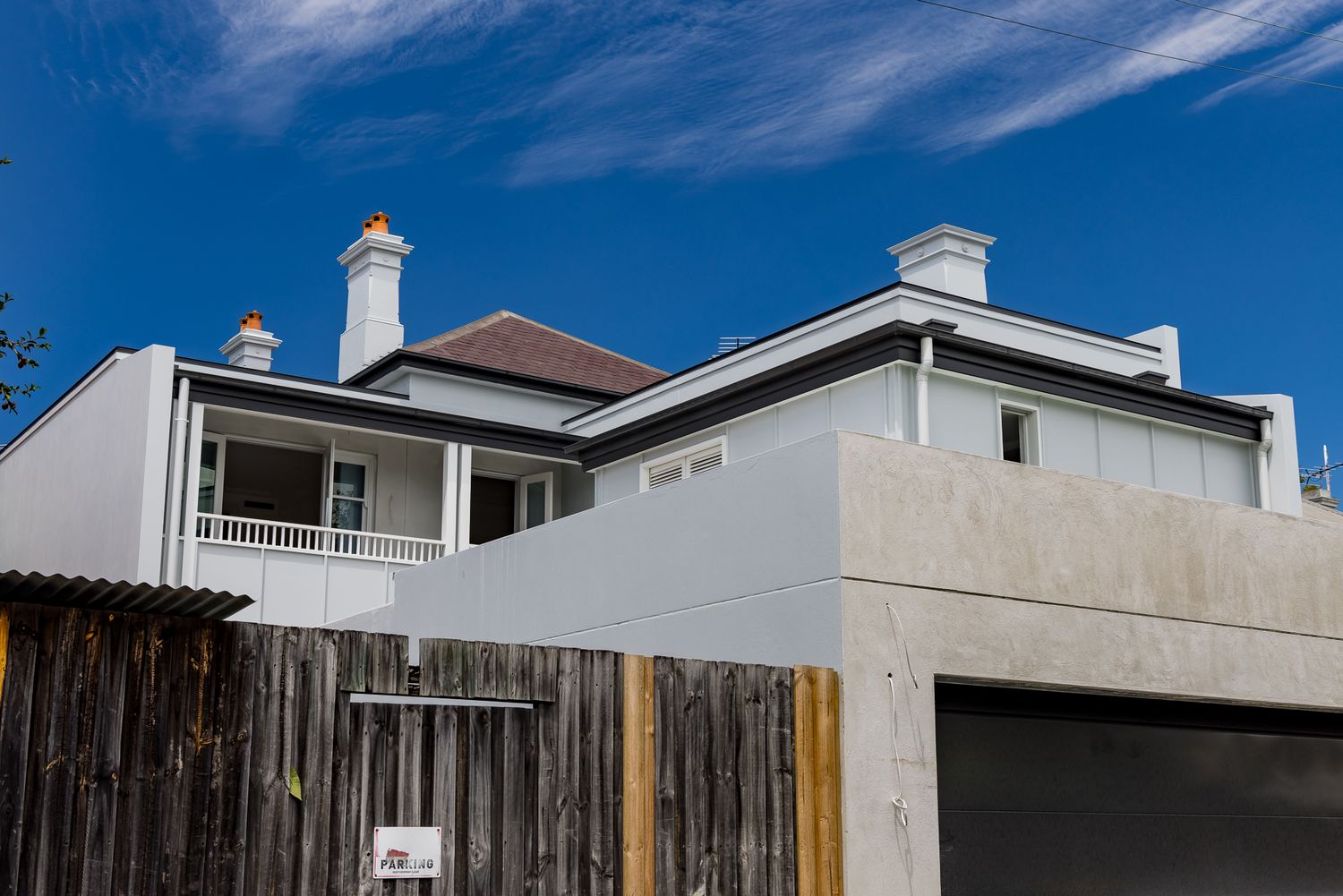 Renovated Victorian-style house with white rendered walls, chimneys and a modern concrete extension beside a timber fence under a blue sky.