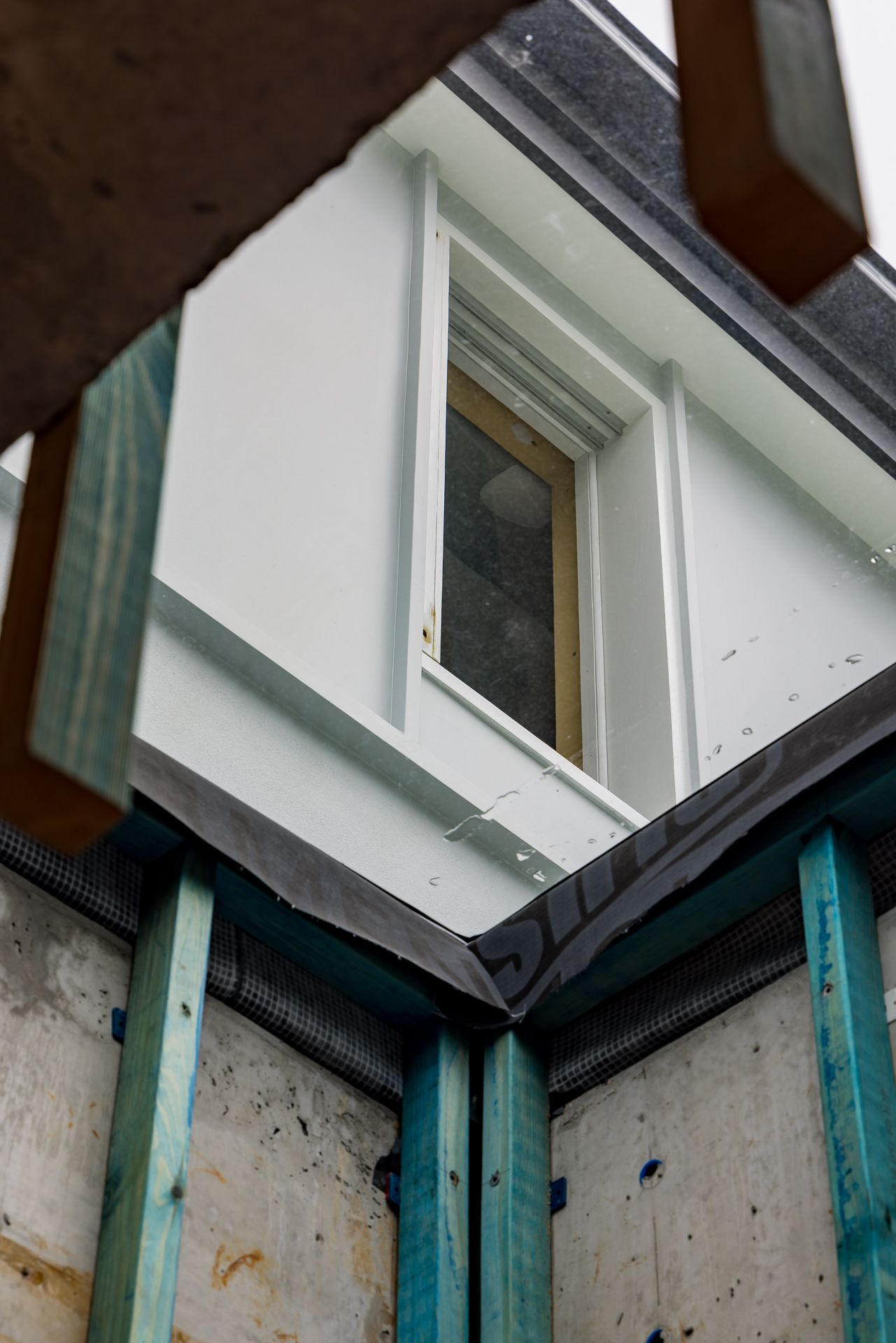 View up to a newly rendered white exterior wall with a timber-framed window above blue timber framing and waterproof membrane.
