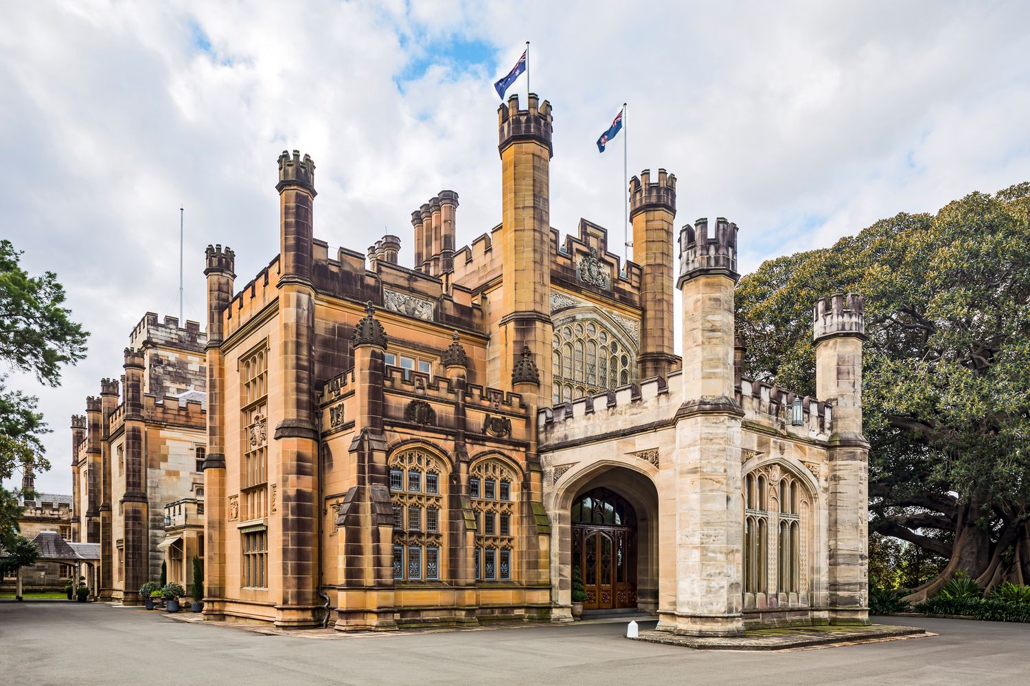 Sandstone Gothic Revival castle with crenellated towers, pointed arched windows, and flags flying beneath a cloudy sky.