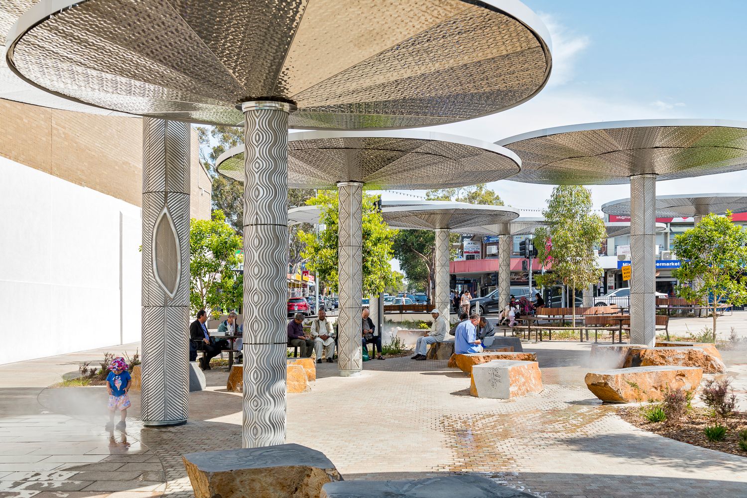 Perforated metal canopies on engraved columns shelter a paved public square where visitors sit among native plants and boulders.