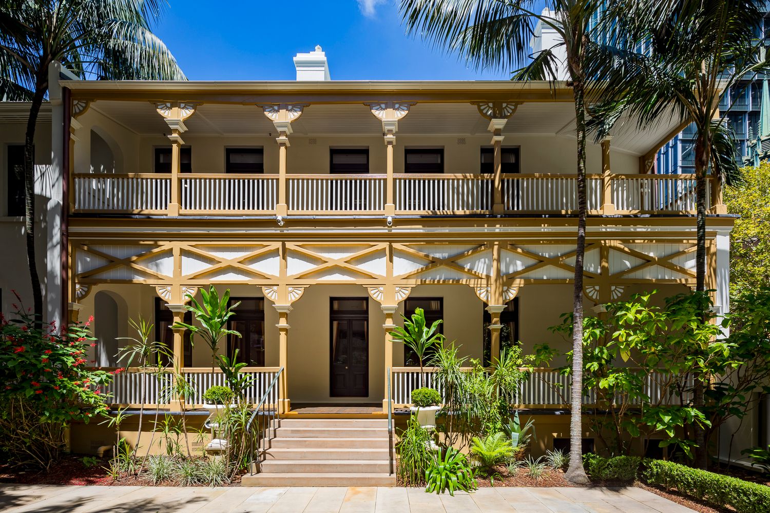 Two-storey heritage building with decorative timber verandah, ornate columns and a staircase framed by tall palm trees and lush greenery.