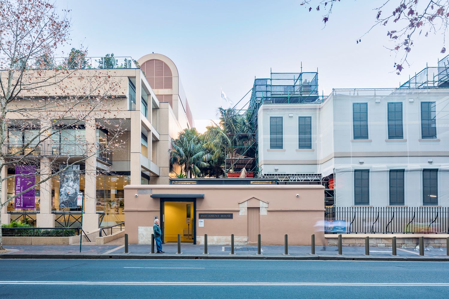 Streetscape of a heritage building under restoration with scaffolding, a modern adjoining building and a glowing yellow entrance doorway.
