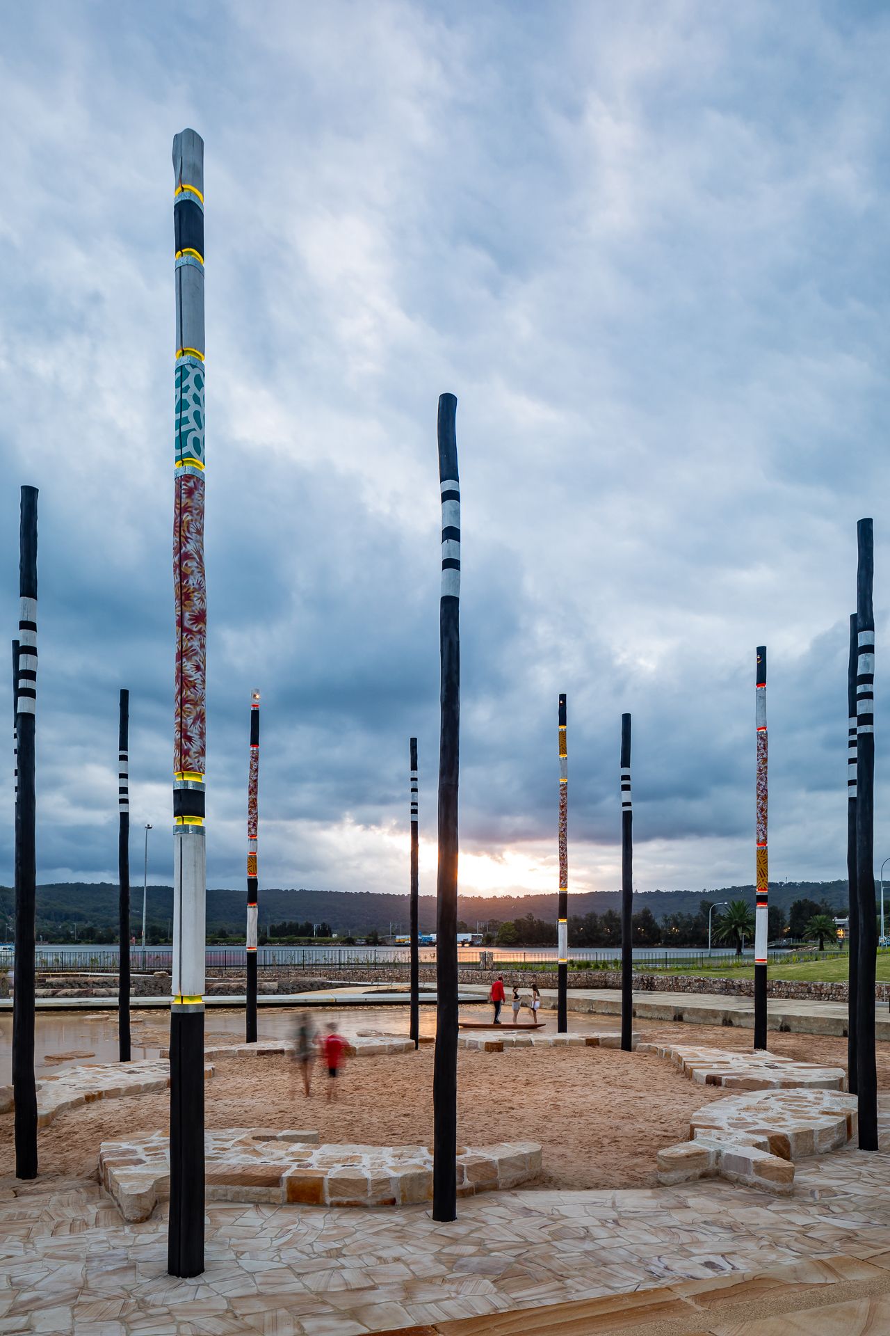 Tall painted art poles encircle a sandy gathering space with stone seating as visitors walk near the water at dusk.