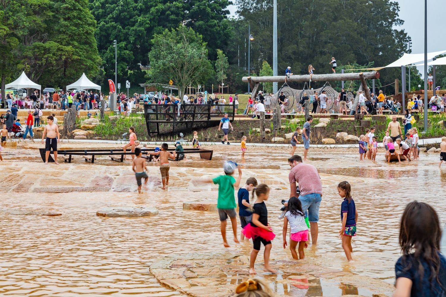 Families and children splash and wade through a shallow sandstone water play area surrounded by trees and market tents.