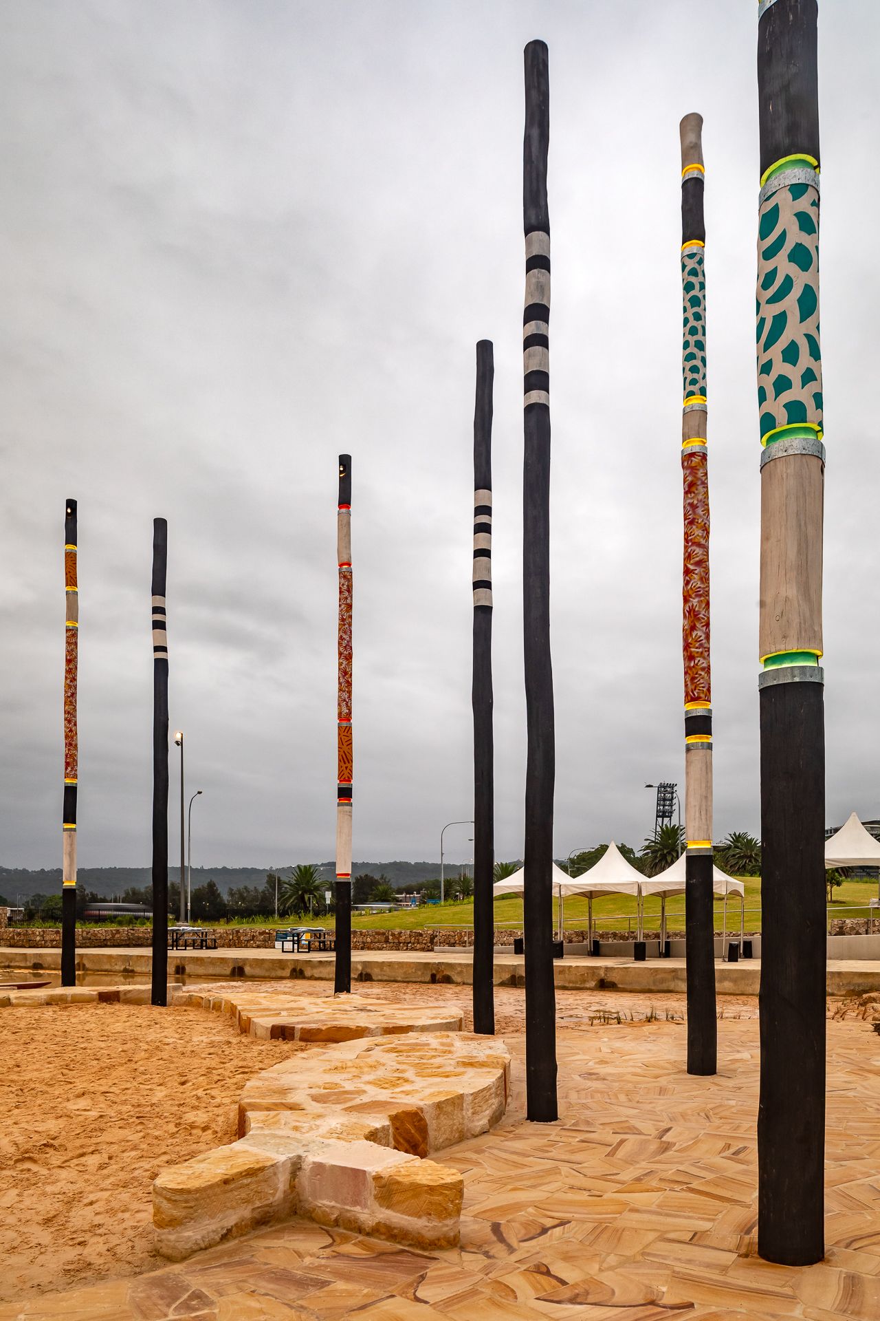 Tall black poles with colorful Aboriginal-style patterns rise from a sandstone and sand plaza under an overcast sky.