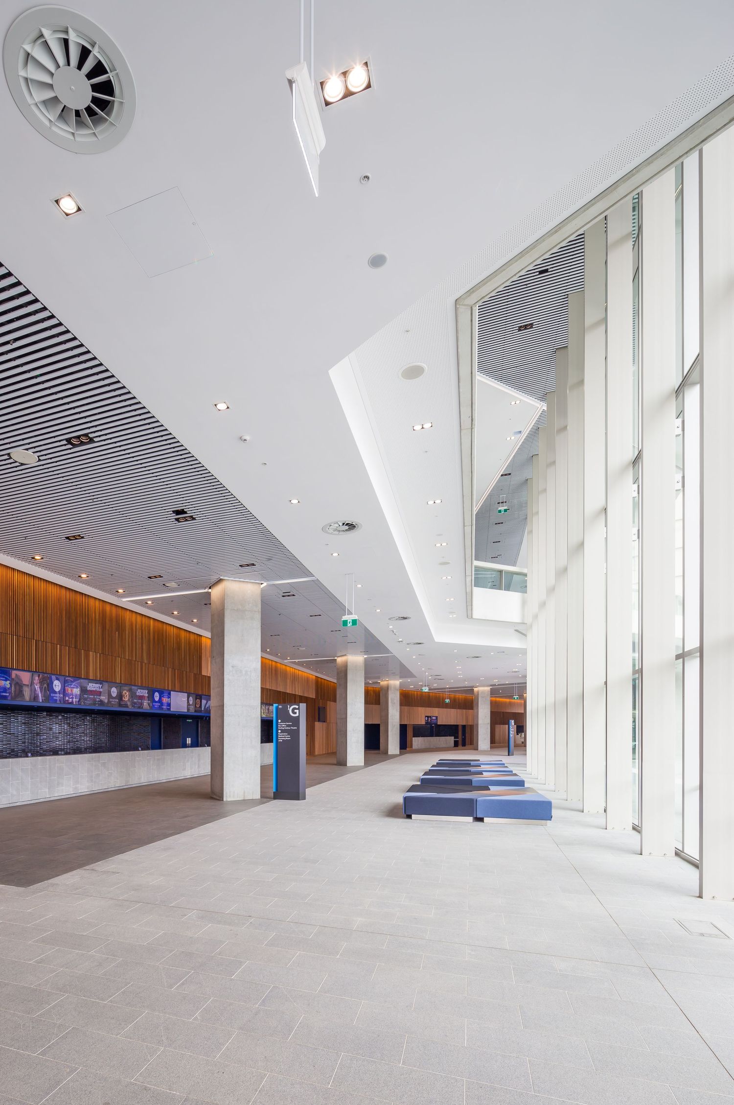 Bright, airy foyer with timber-lined ceiling, tall white columns, floor-to-ceiling windows and blue benches on a stone floor.