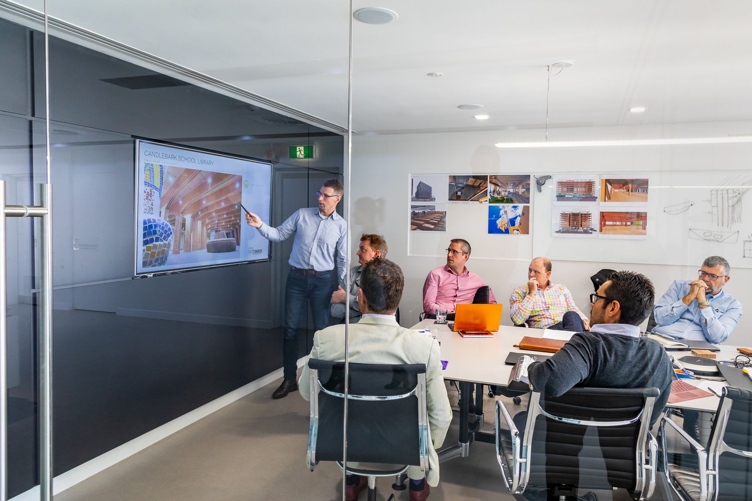 Man presents photos on a wall-mounted screen to colleagues seated around a table in a bright modern meeting room.