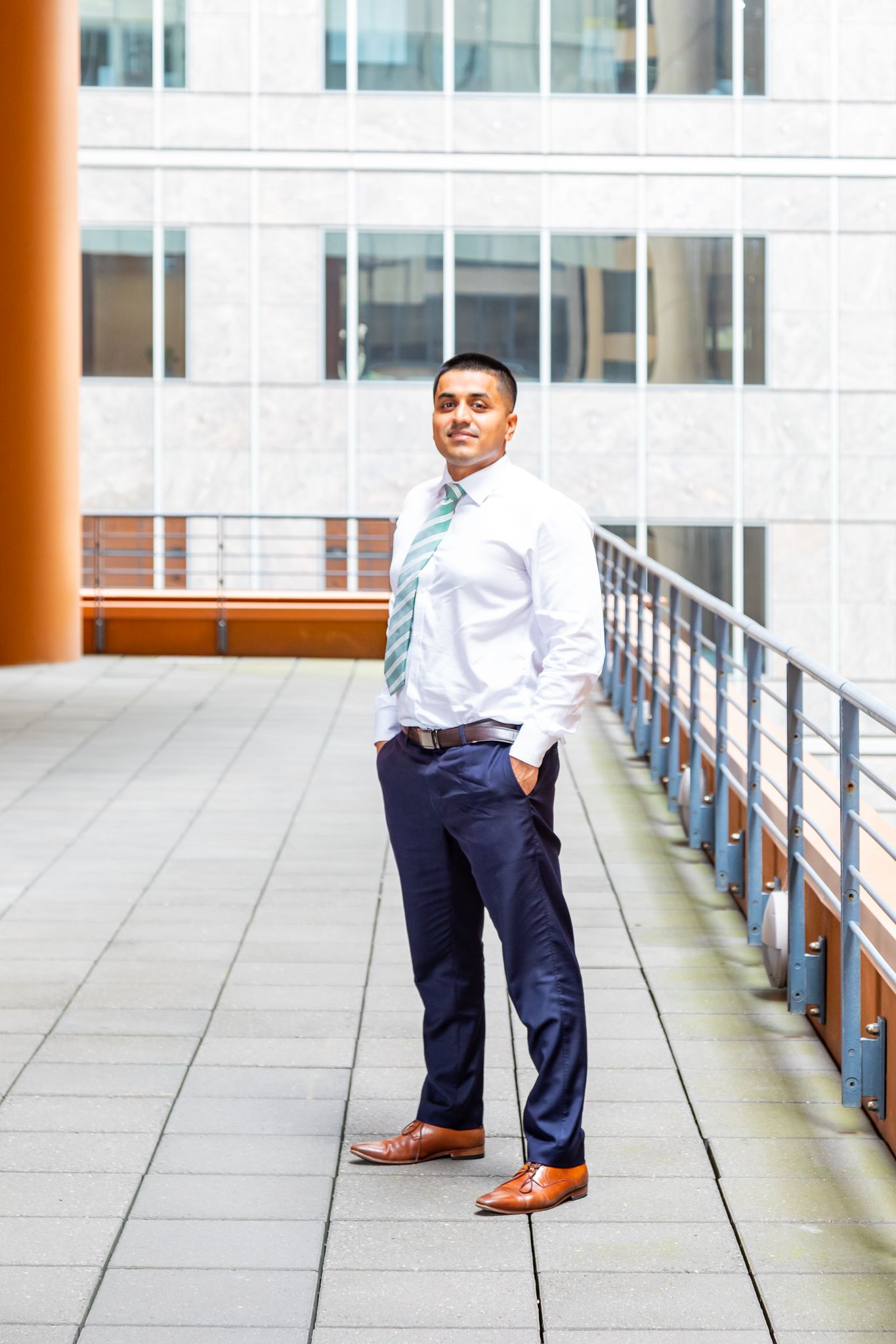Man in a white shirt, green tie and navy trousers poses with a hand in his pocket on an outdoor office terrace with glass building behind.