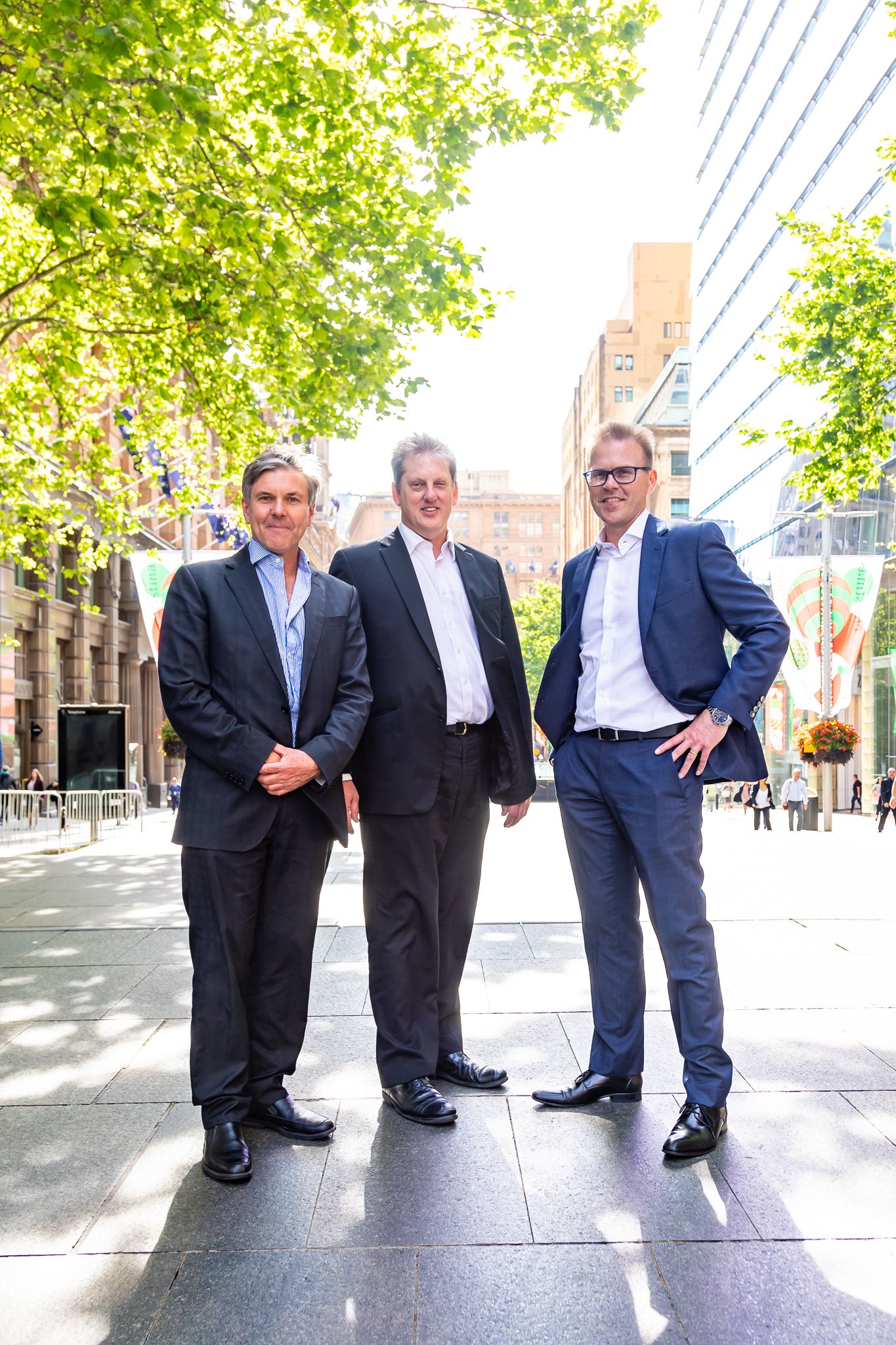 Three men in dark business suits stand together on a sunny city street lined with trees and tall buildings.