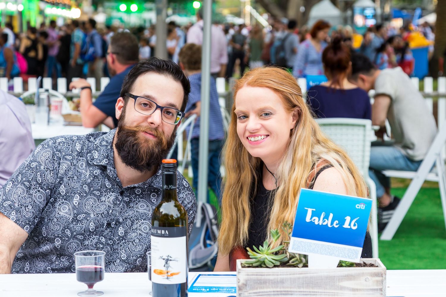 A bearded man in glasses and a smiling woman with long red hair sit at an outdoor event table with a wine bottle and Table 16 sign.