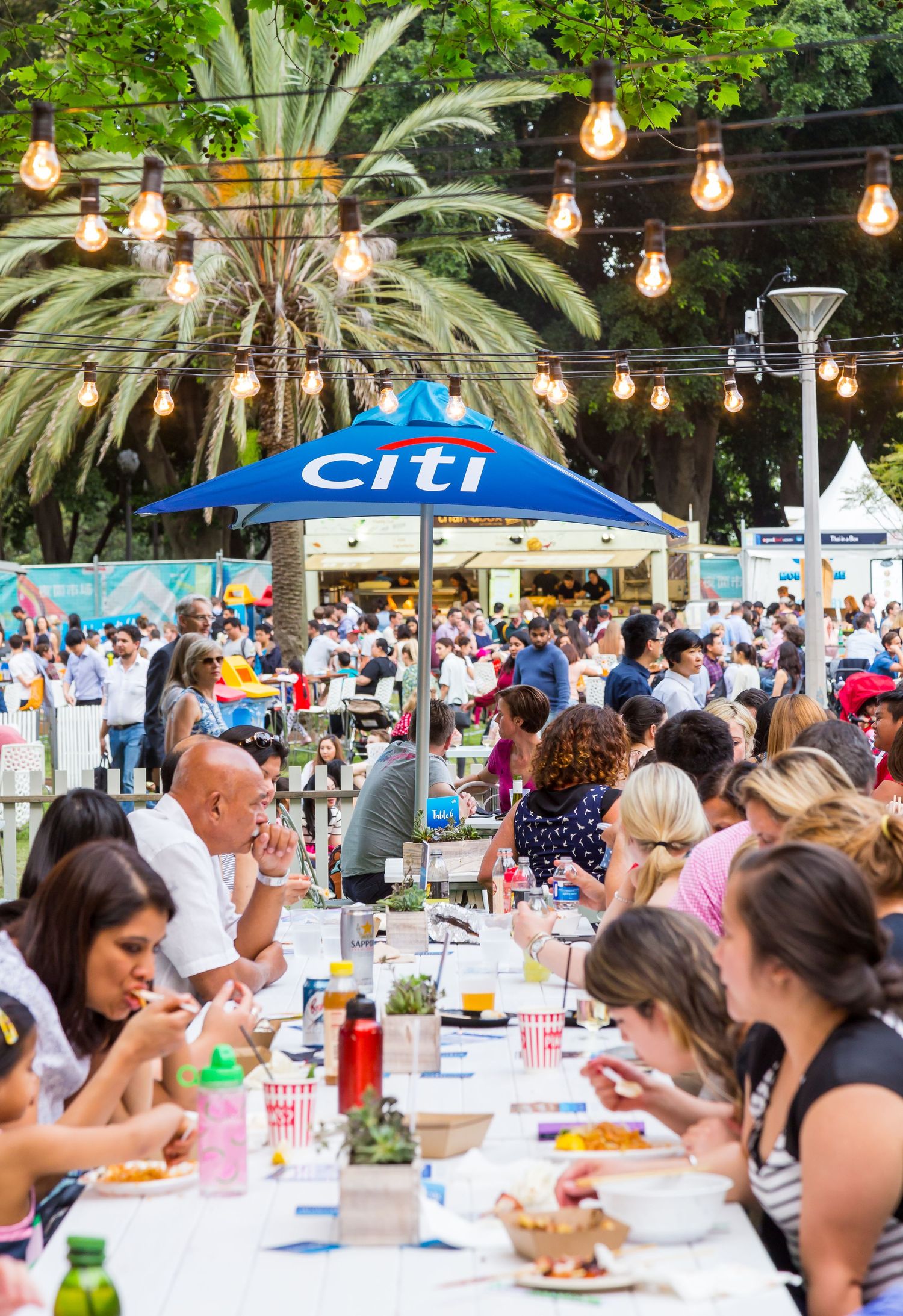 Diners sit at long communal tables under string lights and a blue Citi umbrella at a lively outdoor food festival.