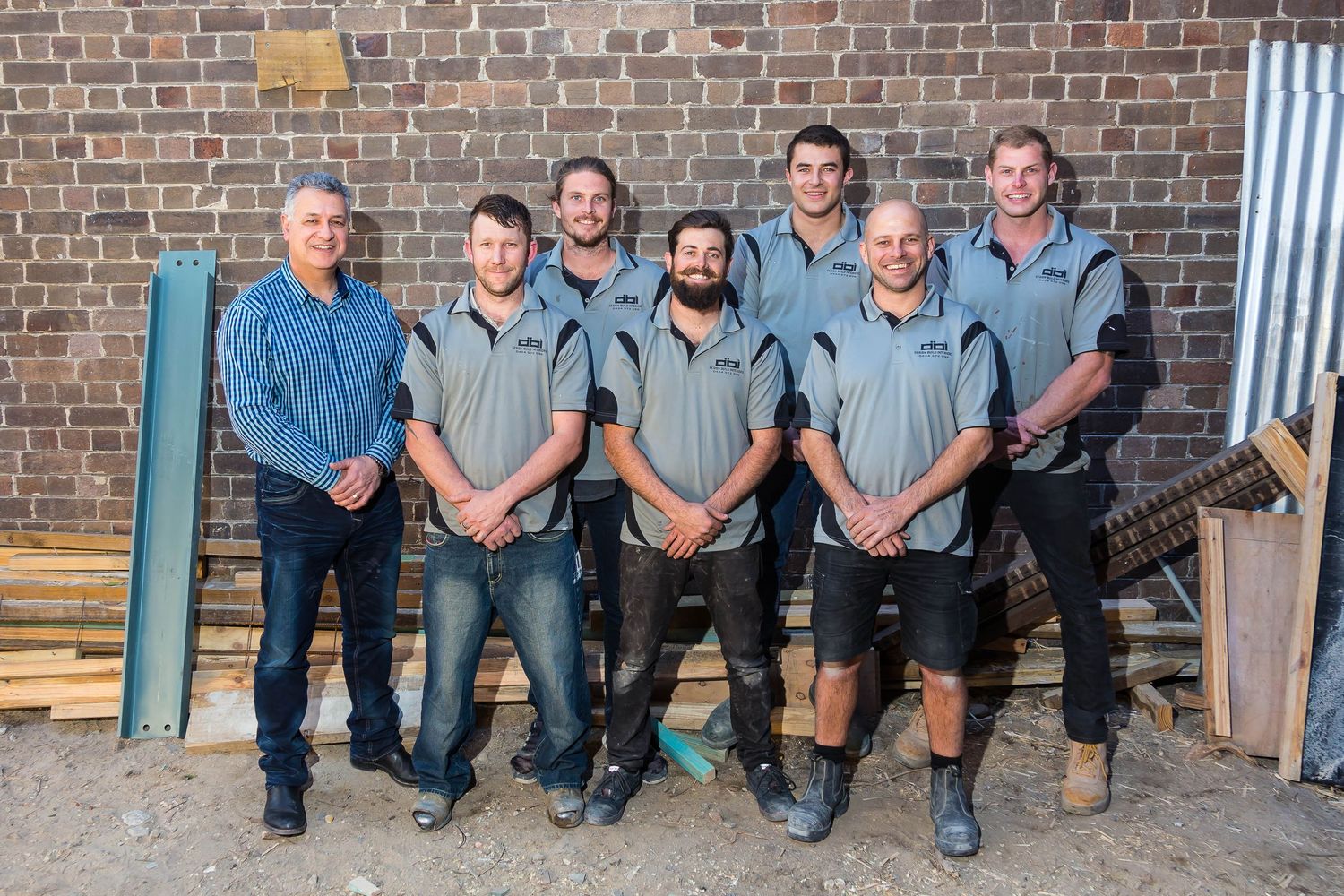 A team of seven men in matching gray work shirts pose together outdoors in front of a brick wall with stacked timber and building materials.
