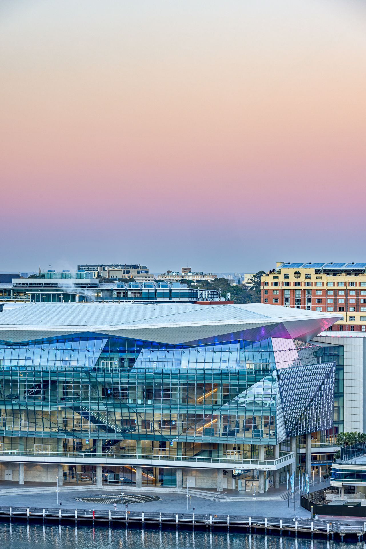 Glass-fronted convention centre at Darling Harbour under a pink and purple twilight sky, with city buildings and the waterfront below.